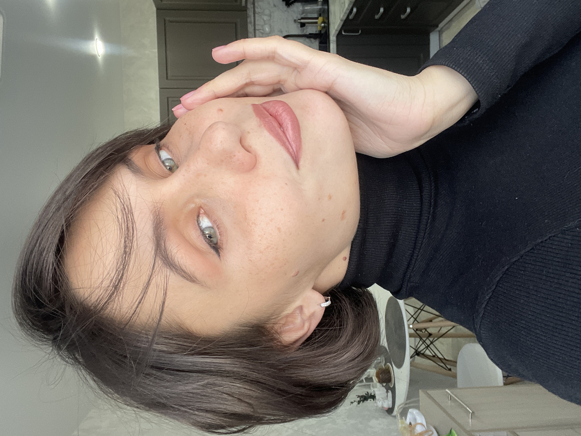 This close-up portrait shows a young woman, likely indoors in Izhevsk, Russia, with her head tilted back and slightly to the right, looking directly at the camera. Her left hand gently rests on her right cheek, showcasing pink polished nails. She has light-colored eyes, possibly grey or green, accented with subtle winged eyeliner. Her dark, medium-length hair frames her face, with some strands falling across her forehead. Her fair skin features numerous freckles across her nose and cheeks, along with a few small moles. Her lips are full, colored with a nude-pink matte lipstick. She wears a black ribbed turtleneck sweater, and a small silver hoop earring with visible stones is seen in her right ear.
The background is softly blurred but reveals elements of an indoor setting, consistent with a home. To the upper left, a light-colored wall features a bright, modern recessed light fixture. On the upper right, dark cabinets or furniture are visible, suggesting a kitchen or dining area. In the lower right, glimpses of light-toned furniture, including a table and parts of a modern chair with a wireframe design, can be seen. The lighting is bright and even, casting soft shadows, and appears to be either natural daylight from a window or well-distributed indoor lighting. The woman's expression is calm and direct. There is no visible text in the image.