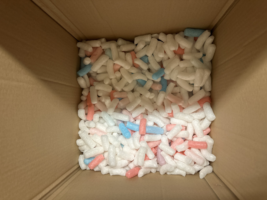 A cardboard box is filled with packing peanuts. The peanuts are primarily white, but there are also light pink and blue pieces mixed in. The packing peanuts are densely packed within the box, filling the space from edge to edge. The box's interior is visible with its brown corrugated texture.