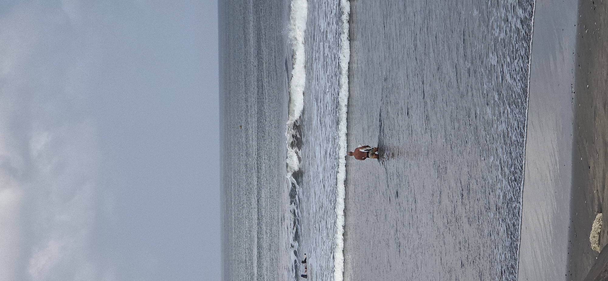 A medium shot captures a lone, bald man wearing dark shorts and holding a surfboard as he stands in knee-deep ocean water. The man faces away from the camera, looking towards the ocean. He is positioned in the center of the frame. In the middle ground of the image, waves crash, creating white foam. Further out, two surfers are visible on their surfboards, appearing to be lying down. The horizon line is clear, with a gray, overcast sky above. The foreground shows a wet sandy beach. The scene is set on the shore of Kuta, Indonesia.