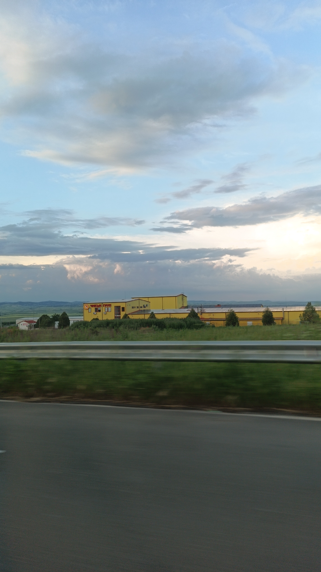 The image shows a roadside view in a somewhat rural or industrial area. The foreground consists of a section of dark gray asphalt road, and slightly blurred, dark green vegetation next to the road.
In the middle distance, there is a long, yellow building with a red sign on it with the words "ЧИСТО ГРУП" in Cyrillic. The building complex appears to be a warehouse or a factory. Some small trees are scattered around the building. In the distance, a flat landscape stretches out towards a horizon dotted with low hills or mountains.
The sky above takes up a significant portion of the image, displaying a blend of blue and grayish-white clouds. The light appears soft, possibly indicating early morning or late afternoon.
The photo appears to have been taken from a moving vehicle. The location is most likely near the city of Burgas, Bulgaria, based on the building's signage.