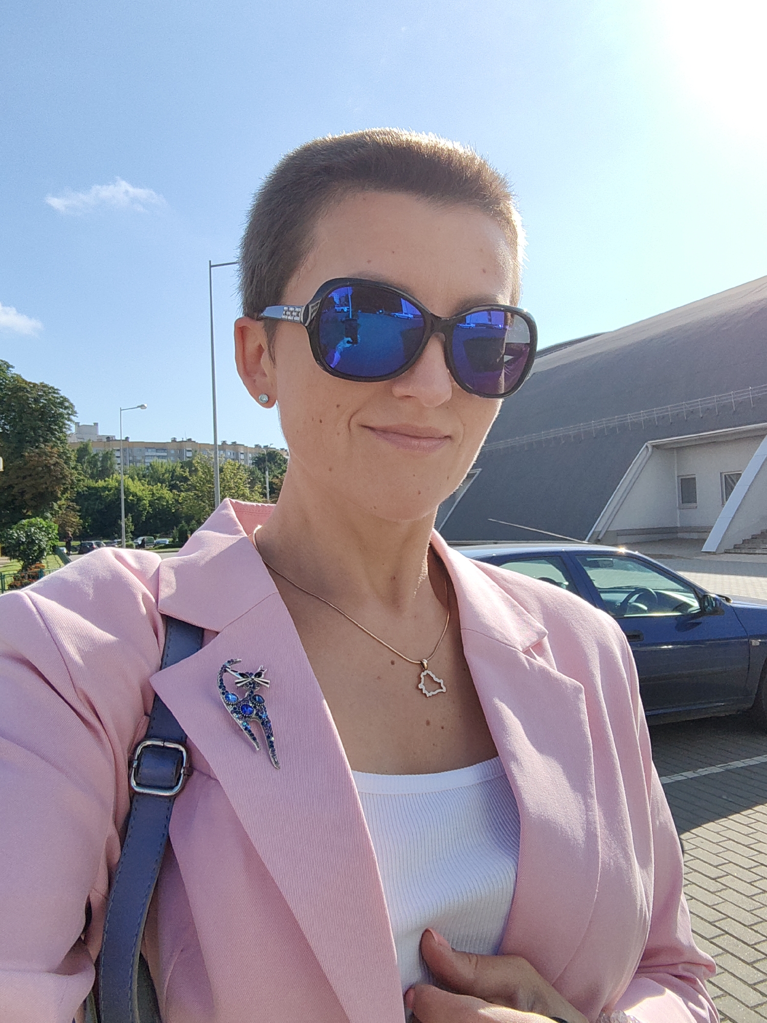 A woman with a short haircut and sunglasses poses for a selfie outdoors on a sunny day. She is wearing a pink blazer over a white tank top and a silver necklace with a pendant. A blue cat-shaped brooch with blue stones is pinned to her lapel. She has a grey purse slung over her shoulder. In the background, there are trees, buildings, a street lamp, a dark blue car, and part of a modern building with a dark curved roof. The ground is paved with light-colored rectangular stones. The woman is standing in an open area, possibly a parking lot or a public square, with a clear blue sky above. Based on the architectural style of the modern building with the curved roof, it is likely the Minsk Arena, a large multi-purpose indoor arena in Minsk, Belarus.