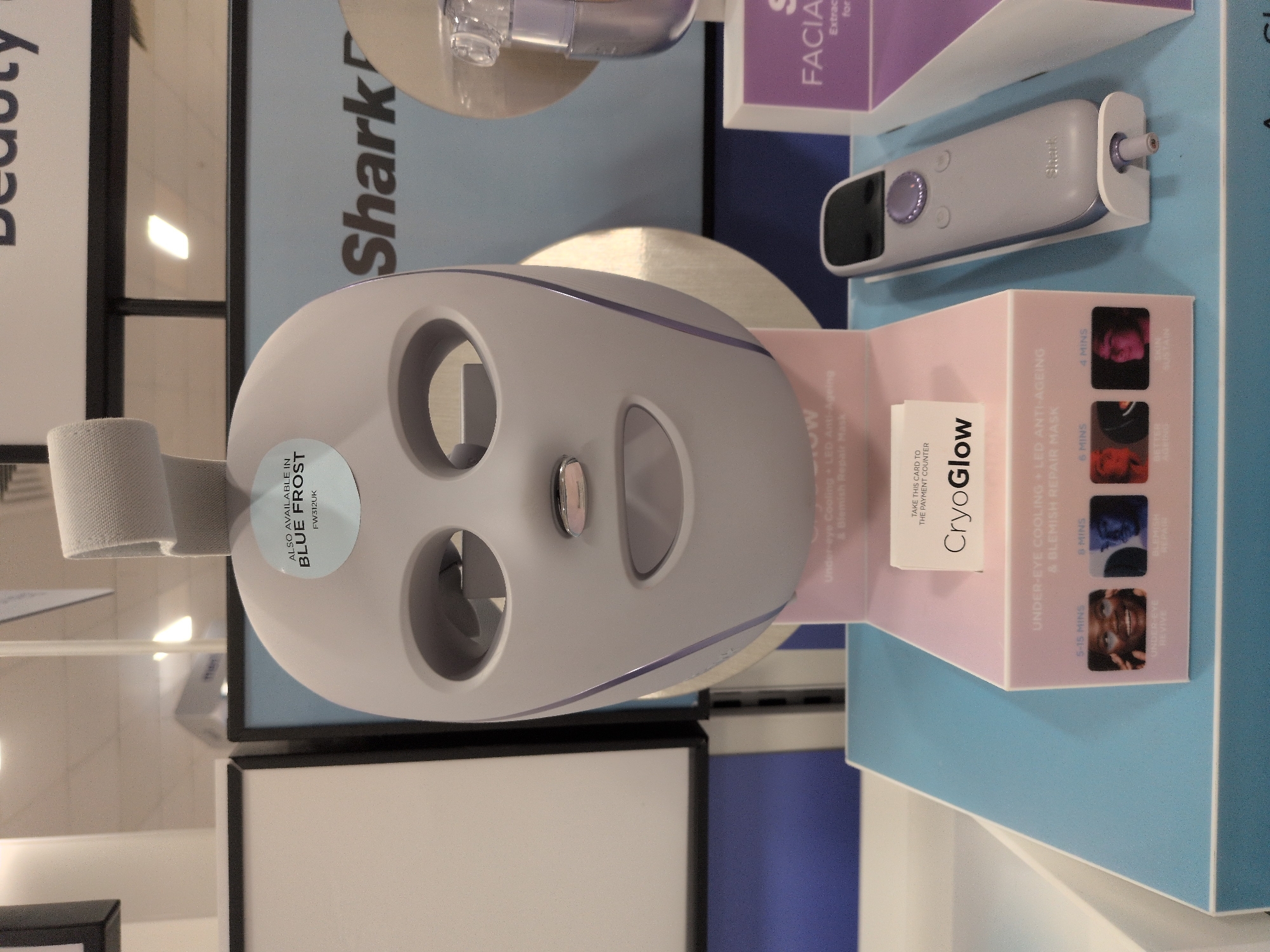 A close-up, eye-level shot captures a beauty device display, likely in a retail setting in Telford, United Kingdom. The primary subject is a light therapy mask, displayed prominently on a pink and white stand. The mask is a pale grey, futuristic design with eye and mouth openings. A circular sticker on the mask reads "ALSO AVAILABLE IN BLUE FROST FW312UK". Below the mask, a small white card displays the text "TAKE THIS CARD TO THE PAYMENT COUNTER" and "CryoGlow". Underneath this, the stand features text and images indicating different treatment times and benefits, such as "UNDER-EYE REVIVE 5-15 MINS", "BLEMISH REPAIR 8 MINS", and "BETTER AGEING 6 MINS". To the right of the mask, a sleek, grey handheld device with a purple dial and buttons, branded "Shark", is displayed on a white stand. In the background, a blue and white sign with the word "Shark" is visible. The overall environment appears to be indoors, with lighting from overhead fixtures reflected on surfaces.