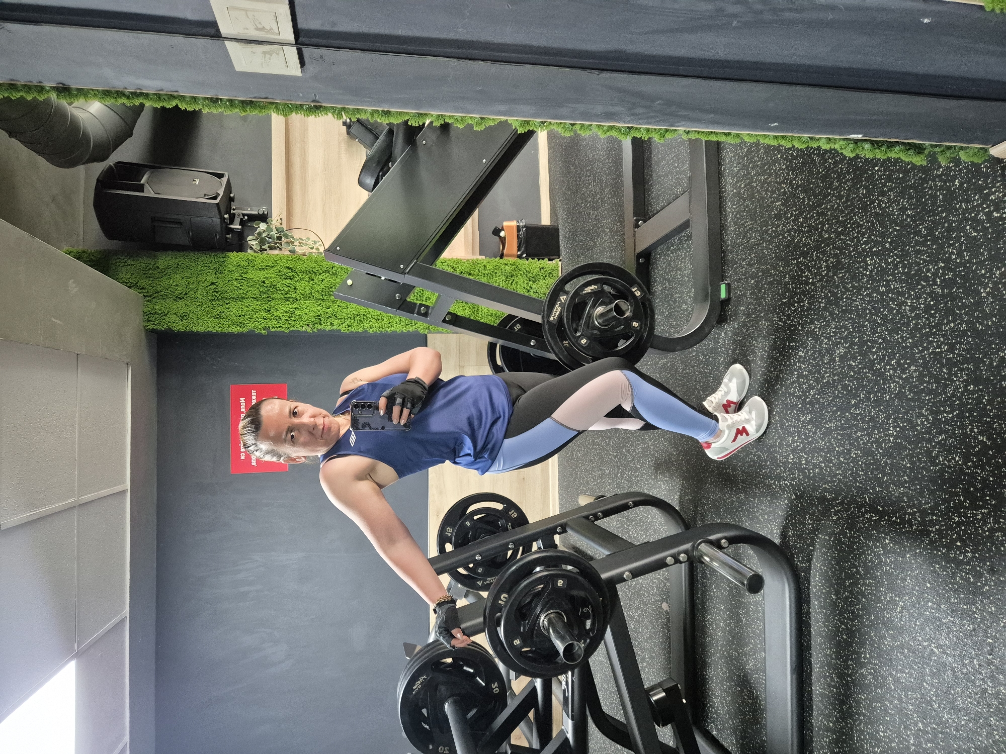 This media file features a woman in a gym, performing a leg press exercise. She is dressed in athletic wear, including a blue tank top, black and light blue leggings, and white sneakers with red accents. The woman is holding a smartphone in her left hand, taking a selfie. She is wearing black gloves on both hands and a bracelet on her left wrist.
The gym setting is evident from the presence of weightlifting equipment, including a leg press machine loaded with weight plates, and a barbell rack with additional plates. The floor is a speckled black rubber mat, common in gyms. Artificial green turf lines some of the walls, adding a unique aesthetic to the space. In the background, a speaker system is visible mounted on the wall, and a large mirror reflects part of the gym. The lighting suggests it is daytime, possibly indoors with artificial lighting. The woman appears focused and determined as she exercises.
A red sign is visible on the wall behind the woman, containing text in a foreign language that appears to be Cyrillic script.