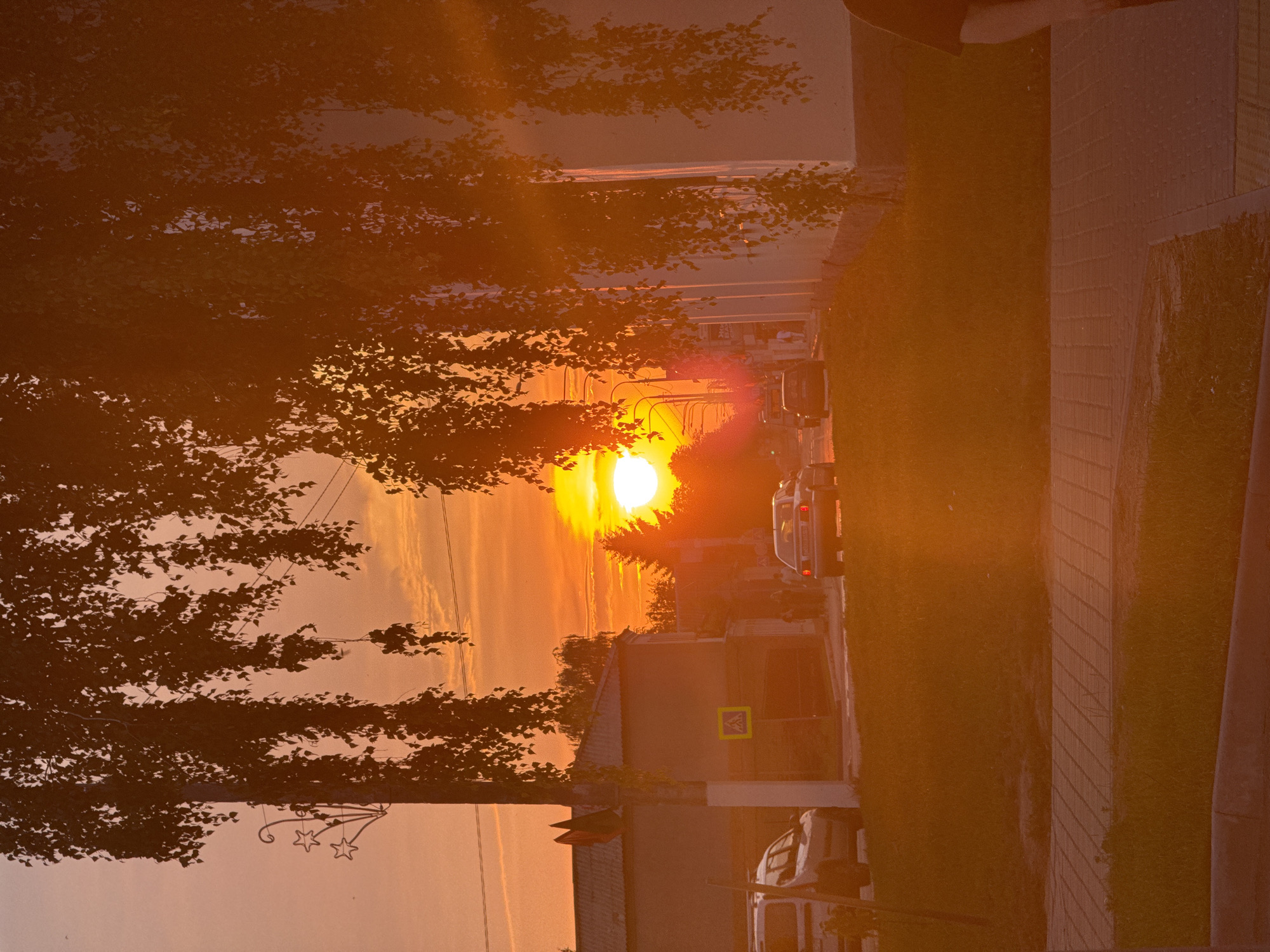 This is an outdoor, daytime, horizontal, medium shot photograph taken at sunset in the city of Nesvizh, Belarus. The sun is setting on the right side of the image, casting a strong orange glow over the entire scene. The sky is bright orange with some clouds. In the foreground, there is a sidewalk with a grassy area. A white car is parked on the left side of the image, and a silver car is driving away from the camera on the right side of the image. There is a building behind the cars. Several people are walking on the sidewalk. There are also trees with hanging branches in the foreground and background.