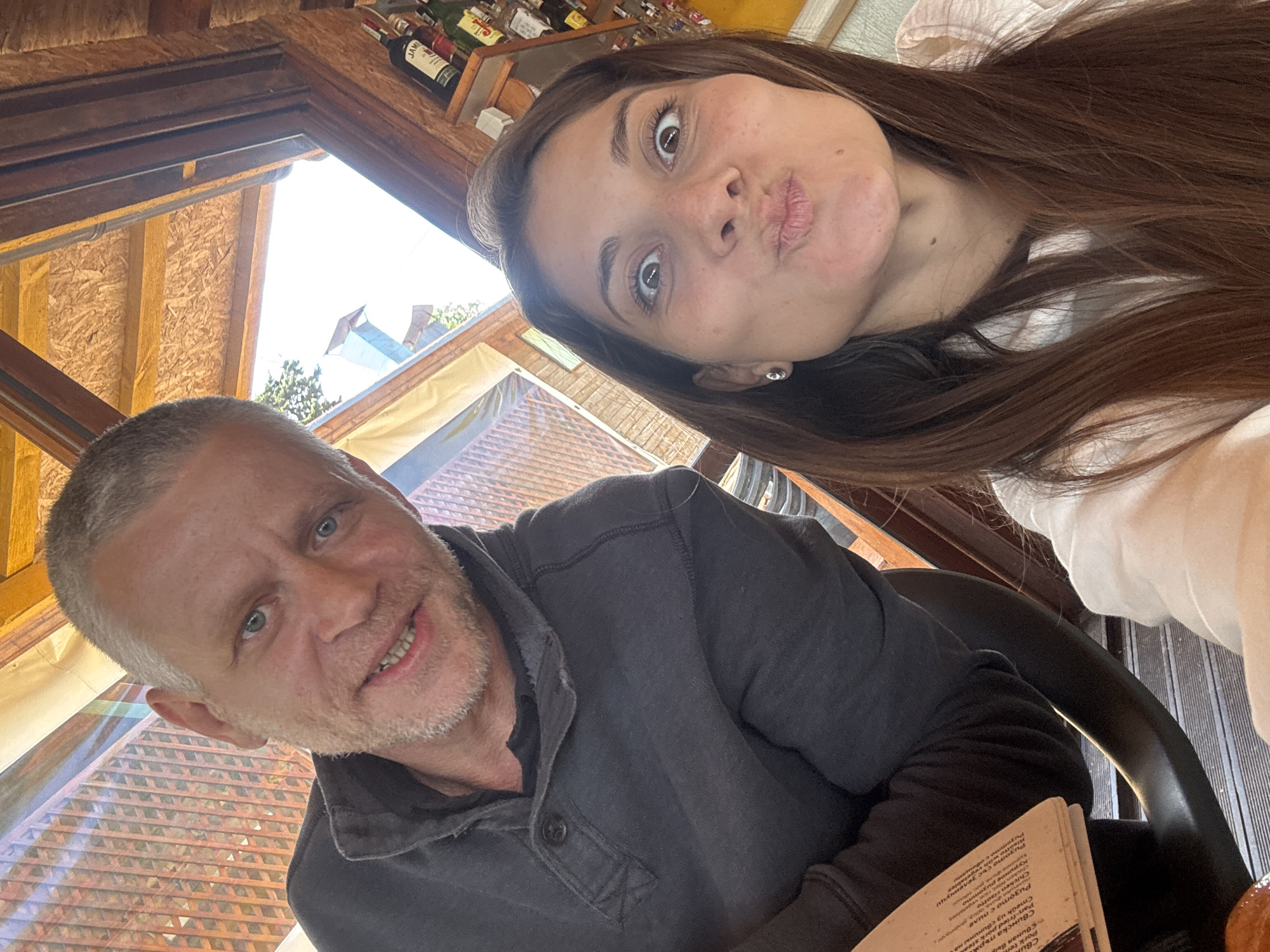This is a selfie taken indoors, likely in a restaurant in Sozopol, Bulgaria, as evidenced by the Cyrillic text on the menu visible in the lower right corner of the frame. A man and a woman are in the shot. The man, who appears older with gray hair, is seated and smiling, wearing a dark gray collared shirt. The woman, with long brown hair, is positioned above and to the right of him. She is making a pouting face for the camera. Bottles of alcohol are visible on a shelf above and behind the woman. A window in the background shows a partial view of the outside with some vegetation. The interior appears to be constructed with wood. A menu is visible in the man's hands.