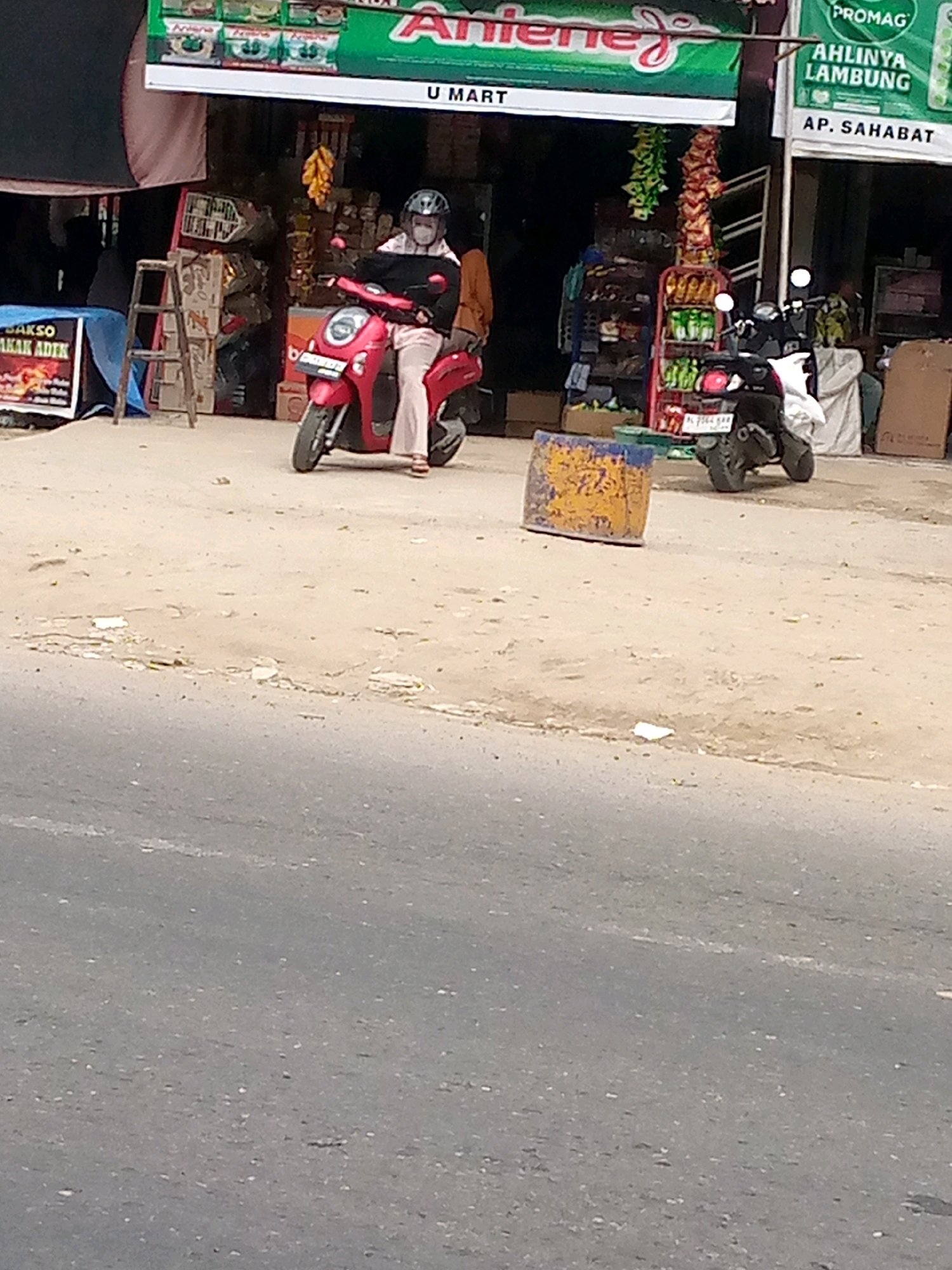 The image depicts a street scene in front of a shop in Keudegeudong, Indonesia. A person wearing a helmet and mask is seated on a red scooter, facing left. The scooter is positioned on a dirt or gravel surface. Behind the scooter, the facade of a shop is visible, with shelves displaying various products. A sign above the shop reads "U MART" and another sign to the right displays "PROMAG AHLINYA LAMBUNG" and "AP. SAHABAT". Two more scooters are parked behind the first one. The environment appears to be an outdoor commercial area.