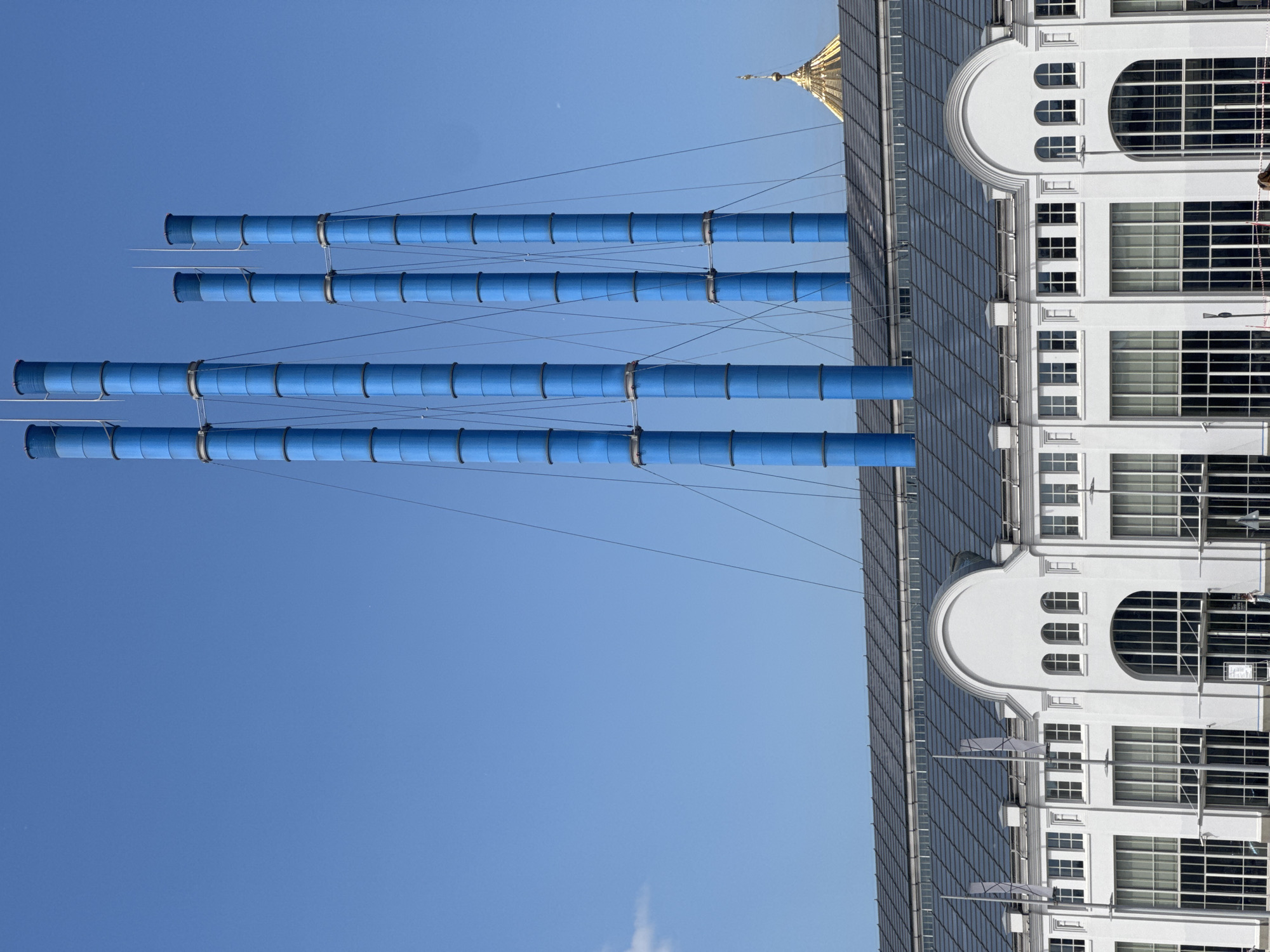 The image, correctly oriented, depicts a clear, bright daytime scene featuring striking architecture against a vivid blue sky. On the left, four prominent, uniformly blue, cylindrical pipes ascend vertically. These pipes appear segmented and are interconnected by thin, taut cables or wires, suggesting they are part of a structural or artistic installation.
To the right of these blue structures, a multi-story white building occupies a significant portion of the frame. Its facade showcases a distinctive curved upper section with three smaller arched windows positioned above a large, semi-circular arched window. Below this, numerous rectangular windows with visible grid patterns punctuate the building's clean, modernist design.
Above the white facade, a dark, steeply sloped roof, possibly made of glass or metal panels, extends towards the right. Peeking from behind and above this roof on the far right, a distinctive golden dome with a slender spire adds a touch of classical grandeur to the composition. The sky is entirely clear and cloudless, indicating sunny weather.
Given the context, the golden dome and the unique blue structures are characteristic architectural elements found within the VDNKh (All-Russian Exhibition Center) complex in Moscow, Russia. There are no people visible, and no specific activities are taking place. No discernible text is present in the image.