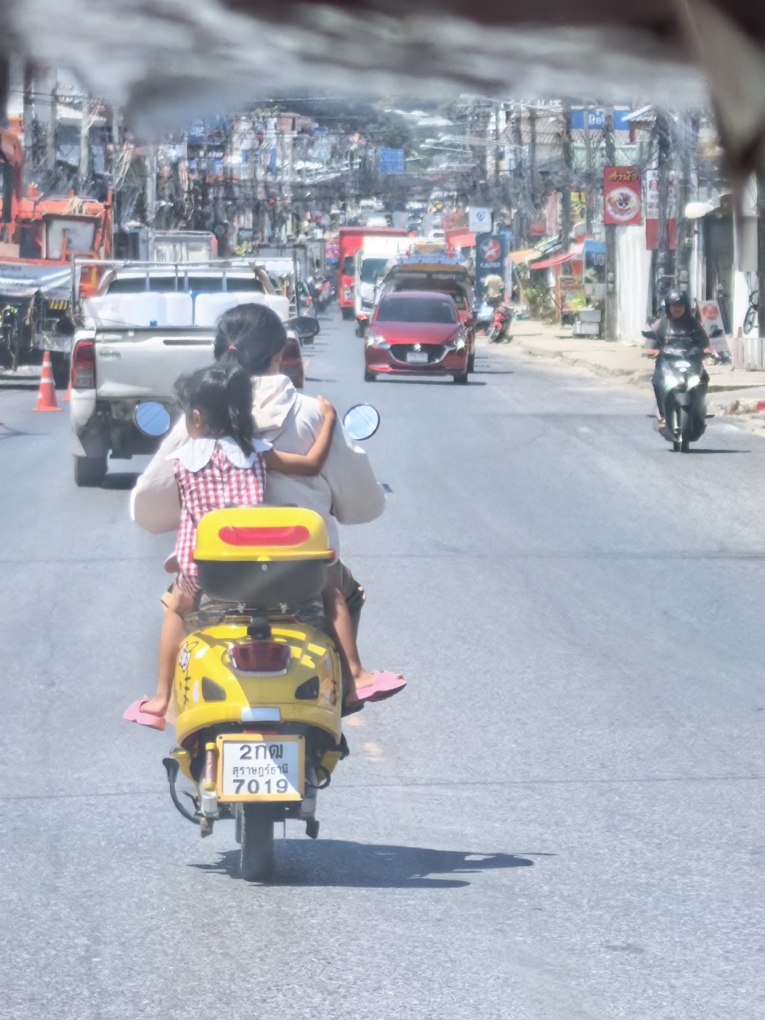 A rear view of two people riding a yellow scooter on a road in the city of Ko Samui District, Thailand. The person in front, likely an adult, is wearing a light-colored shirt and has dark hair tied in a ponytail. They are driving the scooter. Behind them, a child wearing a red and white checkered dress is seated, with their arms around the driver's waist. The child is wearing pink sandals. The scooter has a yellow storage box on the back with red accents and a license plate that reads "2 กม สุราษฎร์ธานี 7019". The road is paved and appears to be busy, with other vehicles visible in the background, including a white pickup truck and a red car. Buildings with signs and a dense network of electrical wires line the sides of the street, indicating an urban environment. The scene is illuminated by bright sunlight, casting shadows on the road.