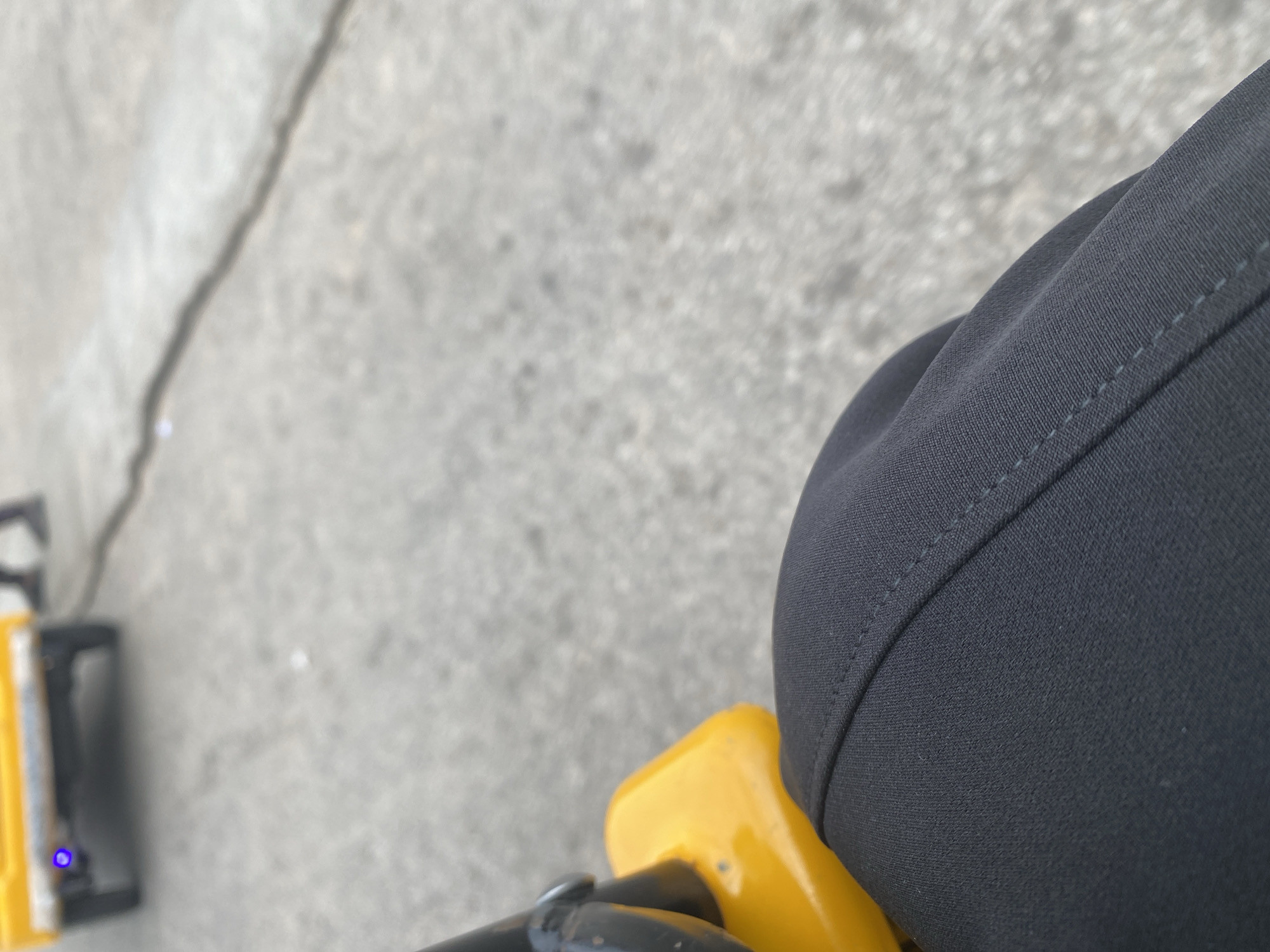 The image is a close-up shot taken from a first-person perspective, likely from someone sitting or operating a machine. The primary subjects are the person's dark-colored clothing, specifically a portion of their leg and thigh, and a bright yellow object that appears to be part of a machine or equipment. The yellow object has a black component and a small blue light is visible on it, possibly indicating it is powered on.
The background is an outdoor scene with a cracked concrete surface, suggesting an urban or paved area. The lighting is somewhat diffused, but there are no strong indications of harsh sunlight or specific weather conditions. The time of day is not clearly discernible.
The overall impression is one of being in the midst of some activity that involves this yellow equipment. The focus is on the tactile details of the clothing and the equipment, with the background providing context for the location. No text is visible in the image. The setting, Jalingo, Nigeria, implies a specific geographical context for this scene.