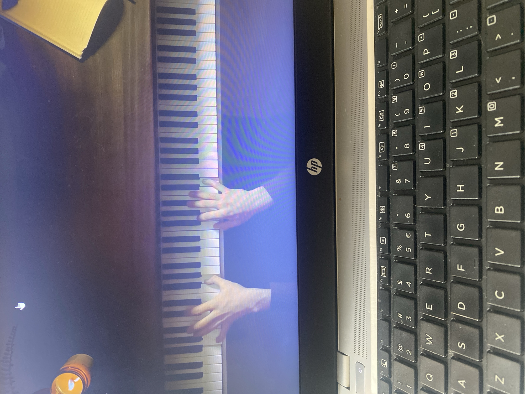 The image displays a laptop screen showing a virtual piano with two hands playing. The keyboard is visible in the foreground. The screen shows a close-up of a person's hands on a piano keyboard, likely playing a virtual instrument on the laptop. The overall scene is focused on the laptop and its display, with no discernible connection to Agadir, Morocco.