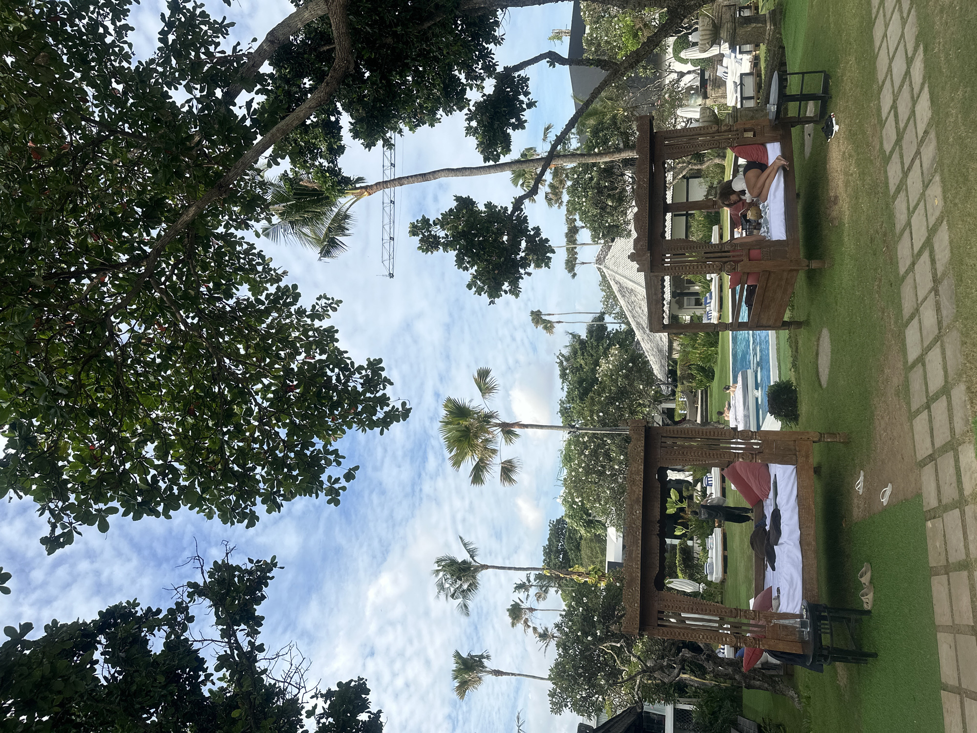 The image depicts a daytime scene in North Kuta, Indonesia, showcasing a tropical outdoor setting. The composition includes tall palm trees reaching towards a cloudy sky, and lush green foliage from various trees and plants surrounding the area. There are two wooden, gazebo-like structures with white bedding and red pillows, appearing as outdoor daybeds. A person is reclining on one of the daybeds. Nearby, there is a swimming pool with people in it and additional lounge chairs. The foreground features more dense greenery. The image has a slightly rotated perspective.