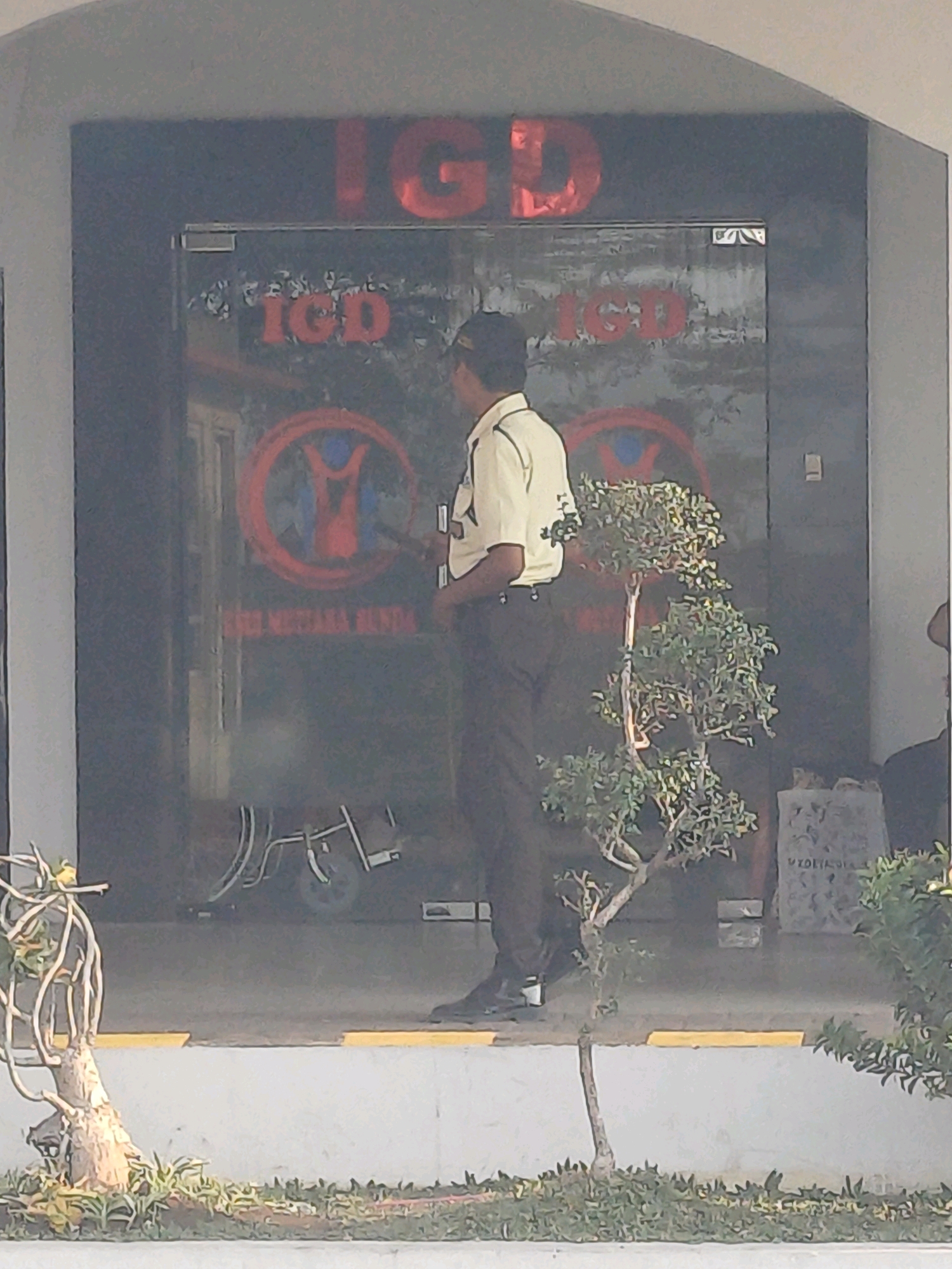The image depicts the entrance to an emergency department (IGD) at a facility in Pejagan, Indonesia. A man, likely a security guard, stands near the entrance. The entrance features large glass doors with the red text "IGD" prominently displayed. Below the "IGD" text, a circular logo with a stylized human figure in red is visible, along with the text "RS. MUTIARA BENDA". A small, decorative bonsai tree is placed to the right of the entrance. In the background, through the glass, a wheelchair or medical cart is partially visible. The environment appears to be an outdoor walkway leading to the building entrance, with planters and landscaping in the foreground and background.