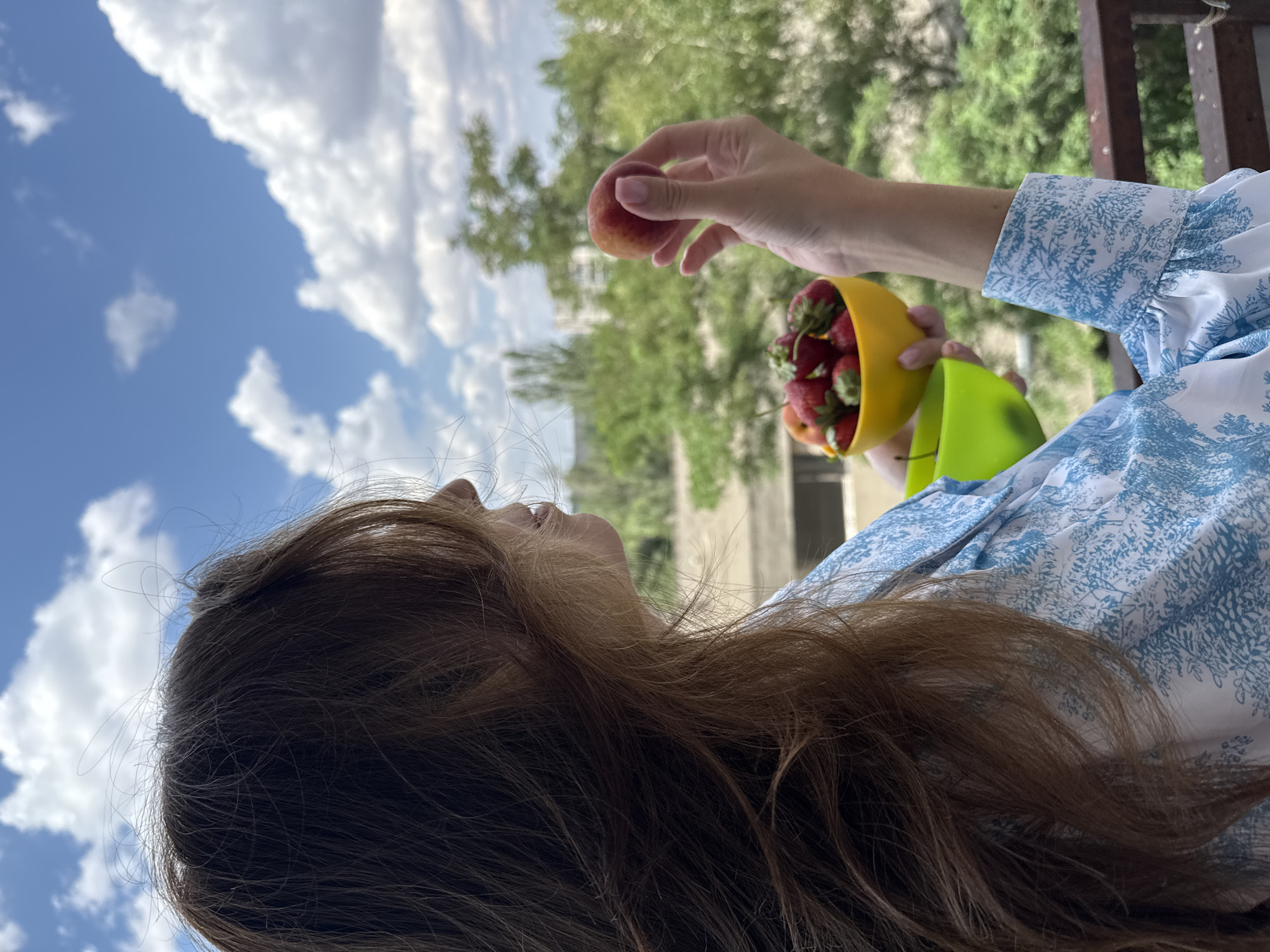 A person with long, brown hair is standing on a balcony in Odesa, Ukraine, holding a yellow bowl filled with strawberries and a peach. The person is wearing a blue and white patterned shirt. The background shows a blurred view of trees, buildings, and a cloudy sky.