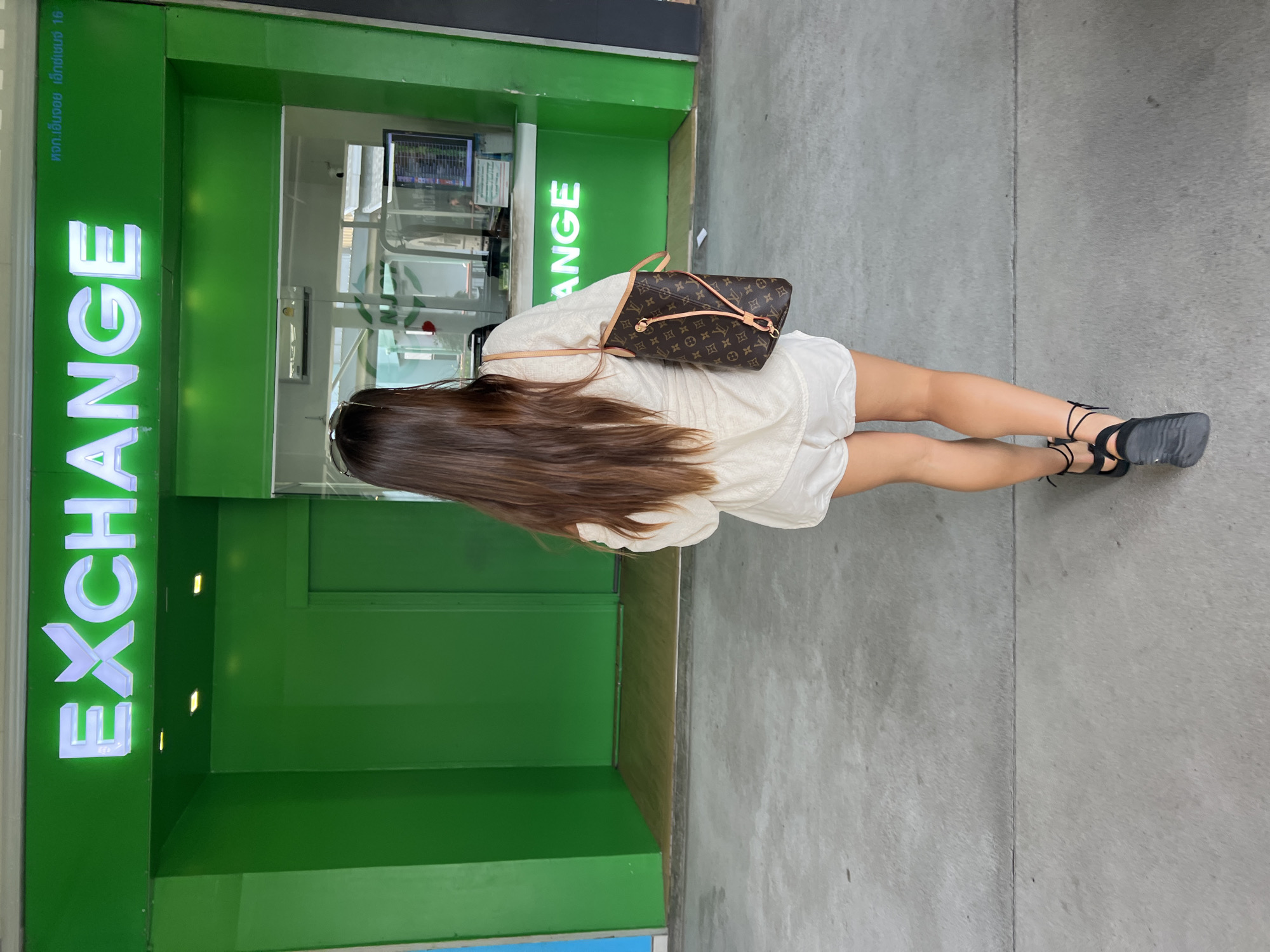 A person walks away from the camera towards a bright green building with "EXCHANGE" displayed in white neon letters. The person is wearing a loose-fitting cream-colored off-the-shoulder top, white shorts, and black strappy sandals. They are carrying a brown Louis Vuitton Speedy bag over their right shoulder. The ground in front of the building is a concrete walkway. The building appears to be a currency exchange booth, with a glass window revealing monitors and a transaction counter inside. The text at the top right of the building, in Thai script, likely indicates the location or branch name. Based on the context provided, this scene is situated in the vicinity of Ban Bang Rak, Thailand.