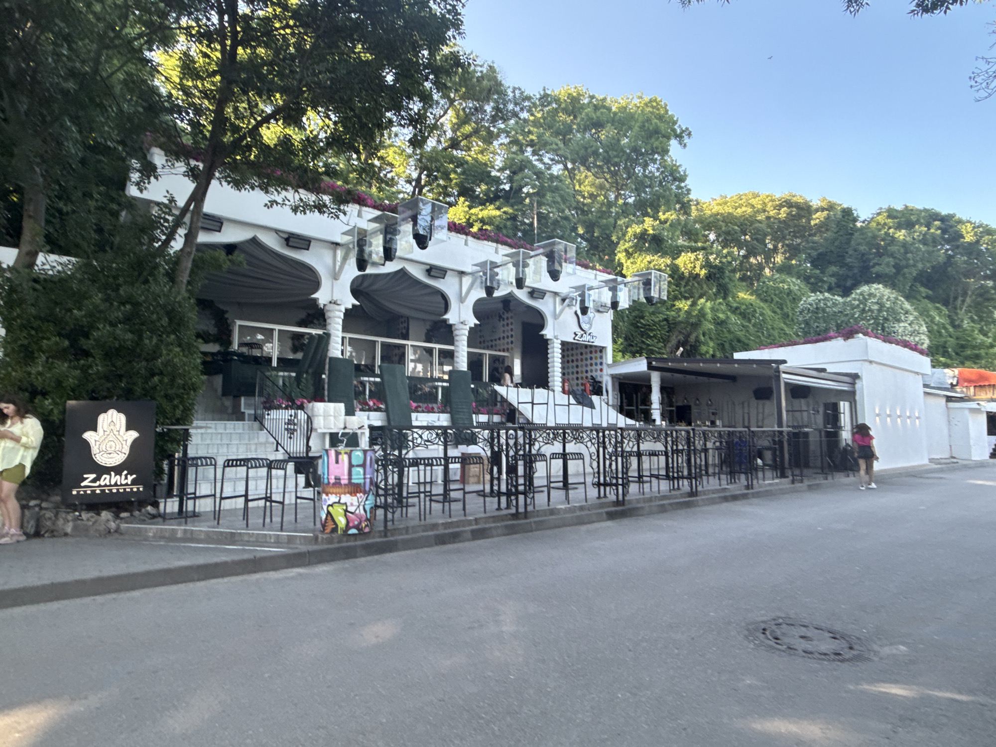 The image shows the exterior of "Zahir Bar & Lounge" located in Varna, Bulgaria. The establishment features a white, multi-tiered building with ornate columns and a large awning. Several outdoor tables and chairs are visible, indicating a seating area. The foreground shows a paved road with a manhole cover. To the left, a black sign with a hand symbol and the words "Zahir BAR & LOUNGE" is displayed. A woman in a yellow top and green shorts is standing near the sign, looking at her phone. Another woman in a pink top and dark skirt is walking away from the building further down the street. Green trees and foliage surround the building, suggesting an outdoor or garden setting. The sky is clear and blue.