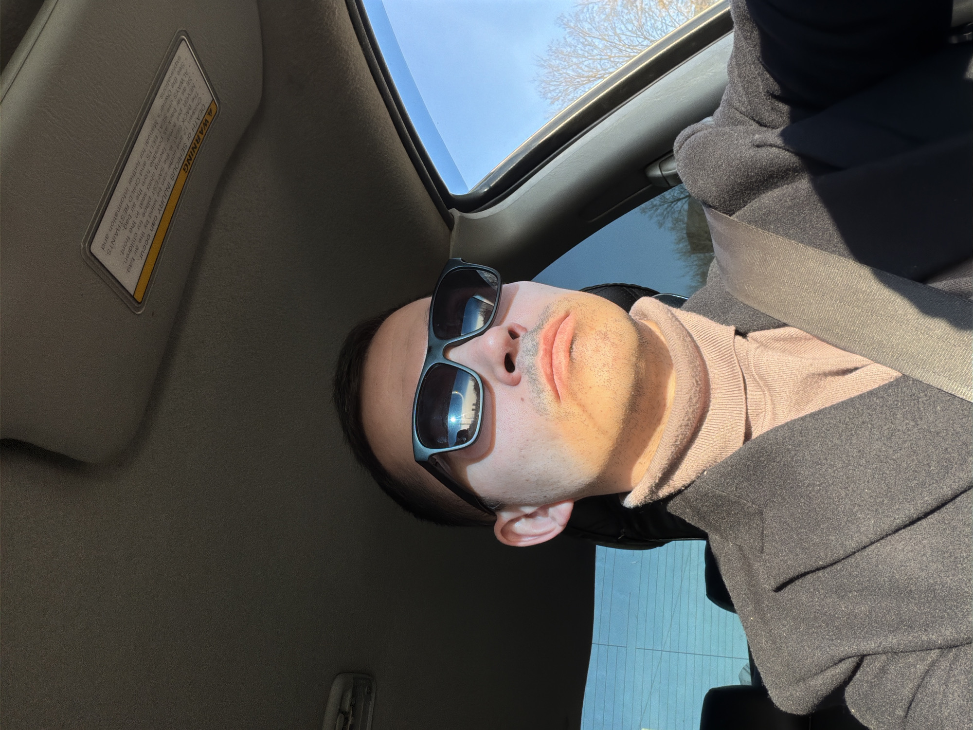 Here is a concise description of the image:
A man wearing sunglasses, a turtleneck sweater, and a dark-colored coat is sitting in the driver's seat of a car. He is wearing a seatbelt. The car has a sunroof. A warning label is visible on the sun visor. The location is likely not Izhevsk, Russia, as the image does not provide any specific information to confirm its location within that city. The environment outside the car appears to be sunny with some trees visible.