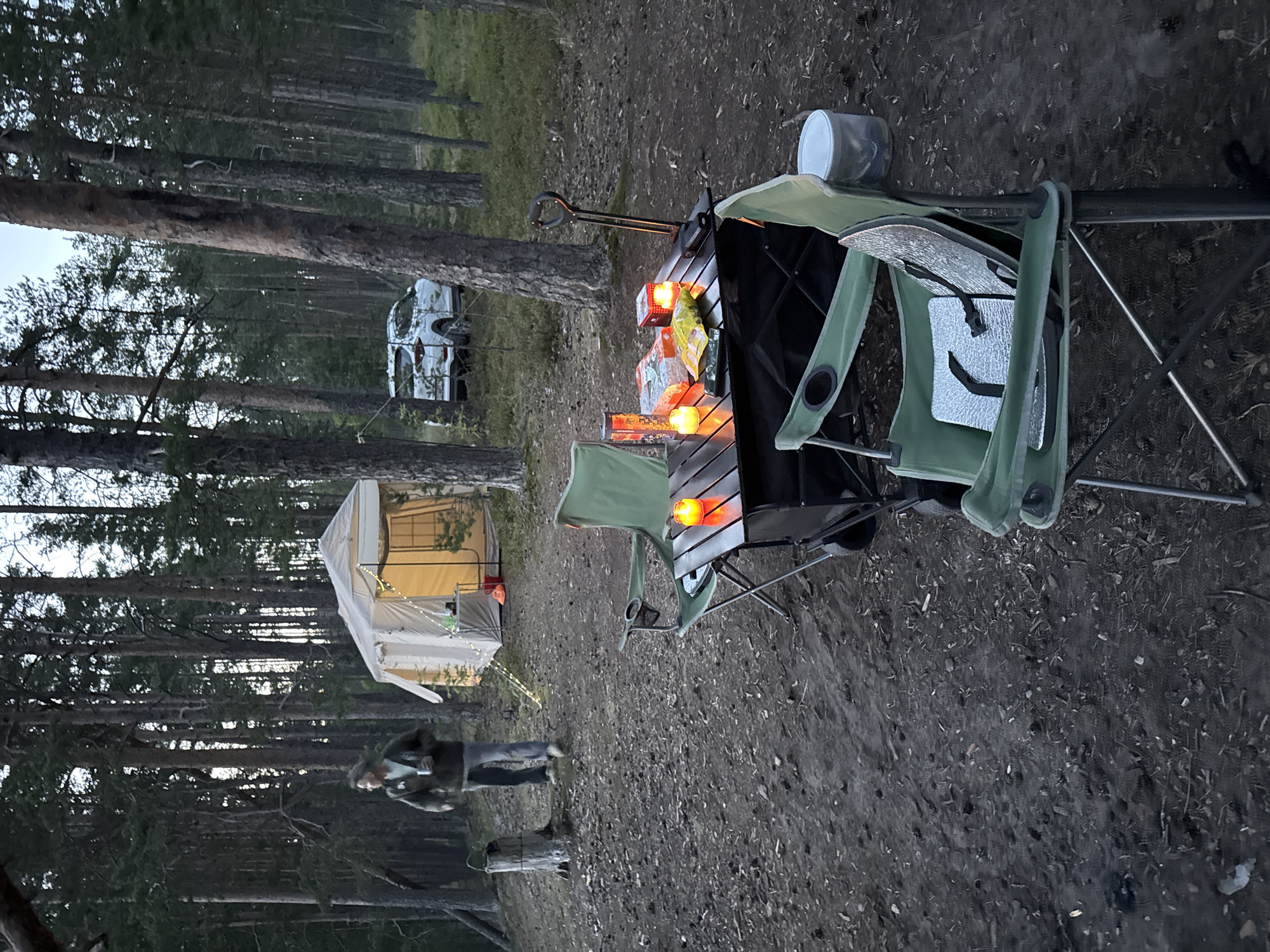In Priladozhskoe, Russia, a campsite is set up in a pine forest. A light-colored canvas tent with glowing interior lights is visible in the background. A white SUV is parked behind the tent. In the foreground, a folding table is set up with two camping chairs. On the table are two lit candles, a package of snacks, and a phone. Another camping chair is positioned to the right of the table. A person wearing a green jacket and jeans walks in the distance, holding a cup. The ground is covered with pine needles and debris.