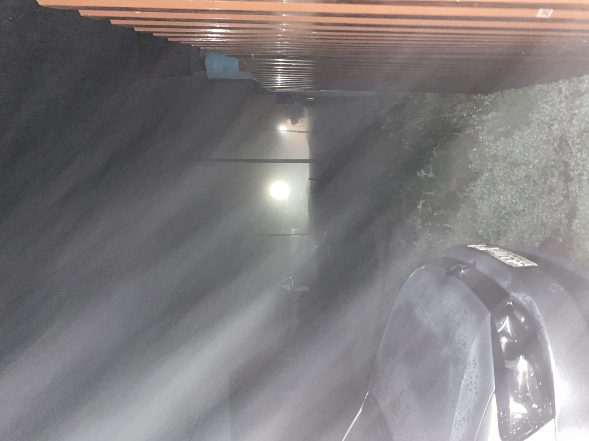 The image is a photograph of a street in Chirsha-Tartysh, Russia, at night. A gray car is parked on the left side of the frame, partially obscuring the foreground. The car's headlights are on, and the hood is wet, suggesting recent rain or dew. In the background, a bright light source, possibly a streetlamp or a vehicle's headlights, illuminates the foggy or misty atmosphere. A tall, thin structure, likely a utility pole, is visible to the left of the light source. To the right of the car, a brown wooden fence runs along the side of the frame, casting a shadow. The ground is covered in low-lying vegetation and gravel. The overall impression is a dimly lit, atmospheric scene in a rural or semi-rural area.