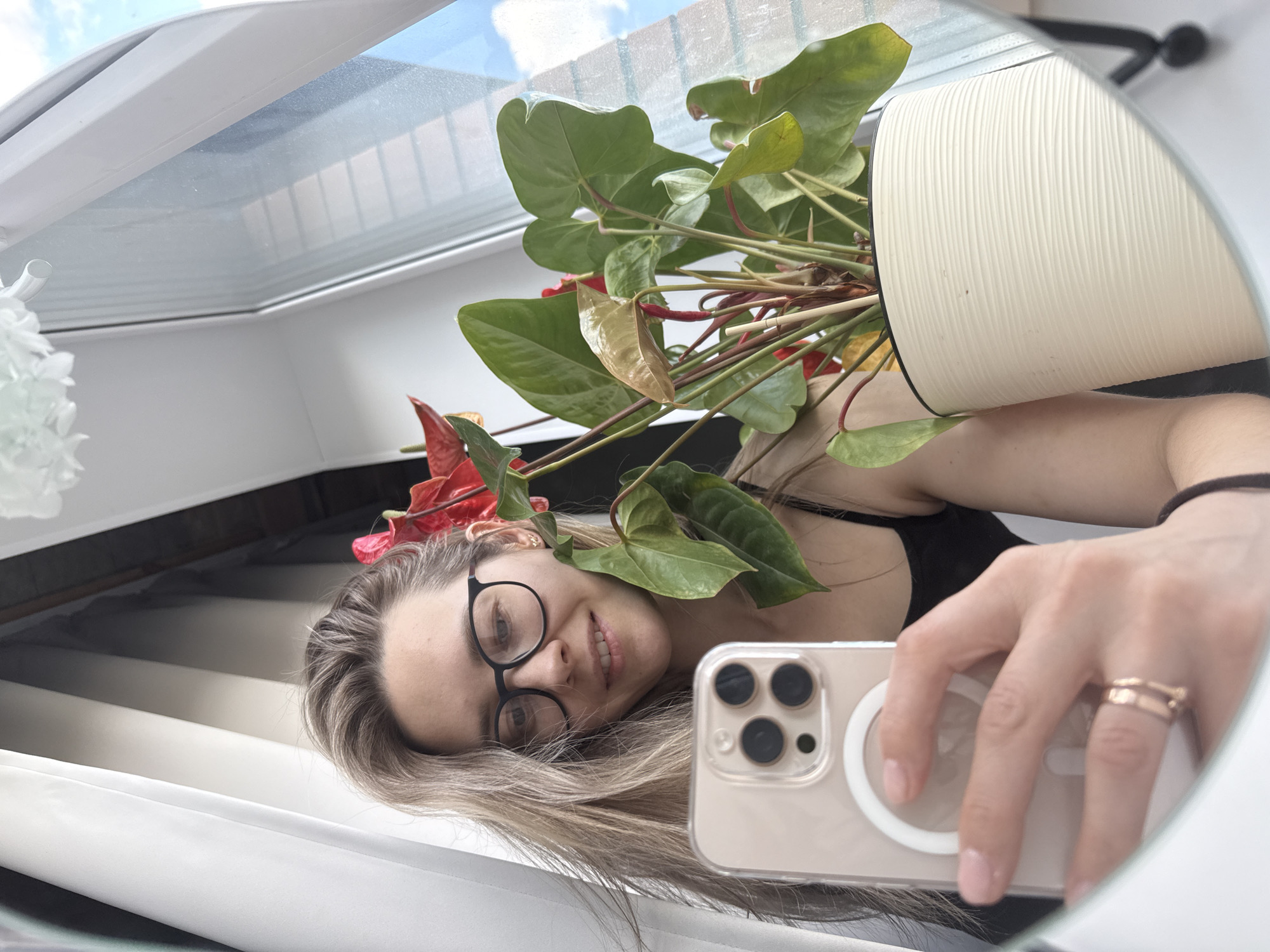 A young woman is captured in a mirror selfie, smiling gently at the camera. She has long, light brown hair and wears black-framed glasses and a black tank top. Her right hand, adorned with gold-colored rings, holds a beige iPhone with a clear case showcasing a MagSafe ring and a triple camera setup.
Behind her reflection, a vibrant potted plant with large green leaves and striking red-pink flowers, resembling an Anthurium, is visible in a white, fluted pot. The background reveals a brightly lit interior space, featuring a large, white-framed window or skylight through which a blue sky with scattered clouds is visible, indicating daytime. The room's aesthetic is clean and modern, with white walls and what appears to be a white ceiling. A partial view of a white, possibly decorative, object is also seen on the left. The overall impression is one of calm and warmth in an indoor setting in Chaikovsky, Russia. No visible text is present.