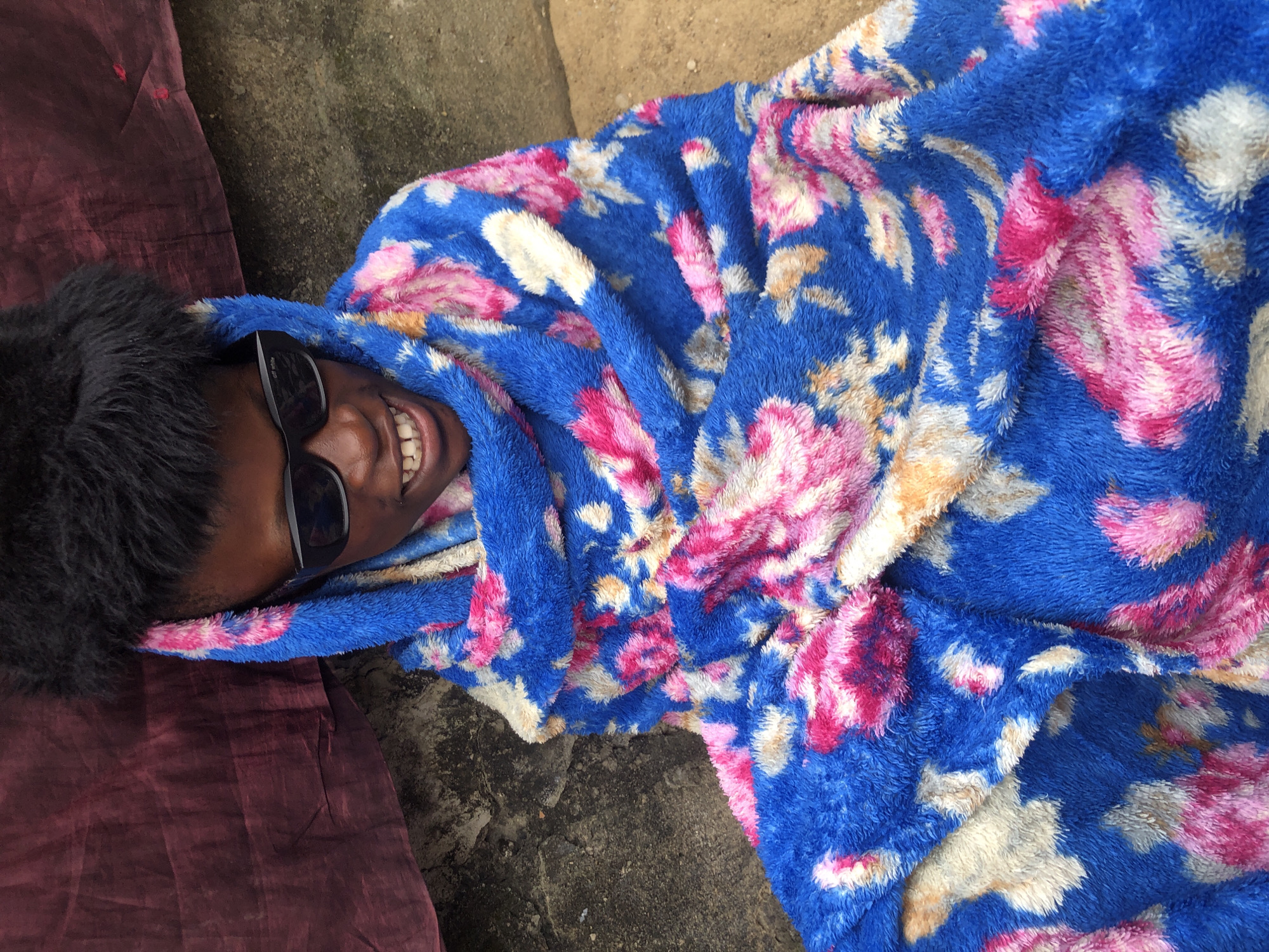 A young person wearing sunglasses and a blue blanket with a floral pattern is sitting on the ground in front of a wall. The person's face is visible, smiling with teeth showing, and their black hair is fluffy. The blanket is wrapped around them, covering most of their body. The background shows a textured wall and the ground, which appears to be dirt or sand. A piece of dark red fabric is draped to the left of the person. The lighting suggests it is daytime, possibly outdoors.
