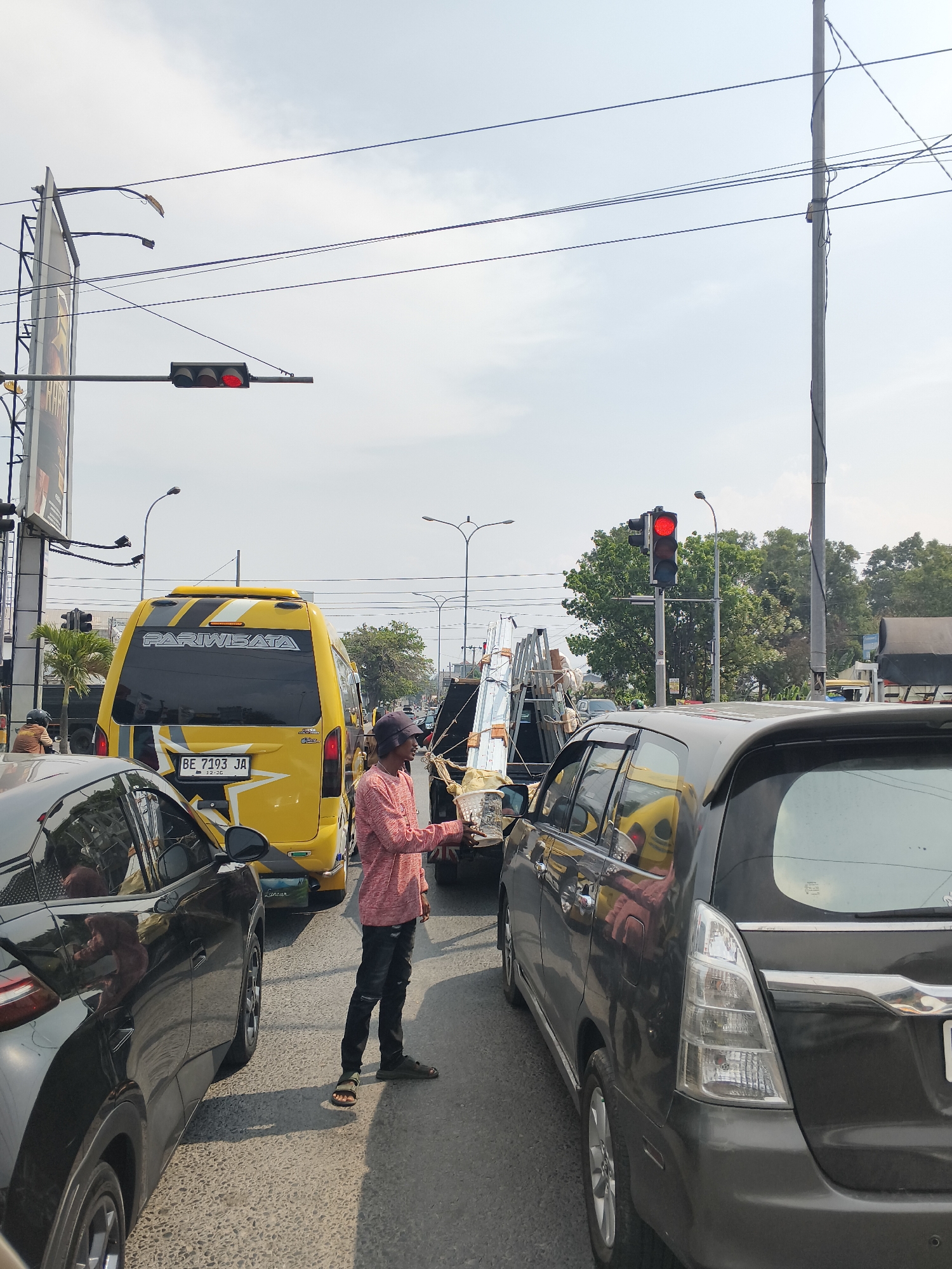 In Bandar Lampung, Indonesia, a traffic scene unfolds at an intersection. A man in a pink long-sleeved shirt and dark pants, wearing sandals, stands on the asphalt road, holding out a white object, possibly a drink or food, to a person in a dark-colored SUV. The SUV is in the foreground, and a yellow bus with the text "PARIWISATA" is to its left. A truck is visible behind the man, carrying stacked metal beams and other materials. Traffic lights are visible, with the light showing red. The sky is overcast. The image captures a moment of commerce or interaction amidst a traffic jam.