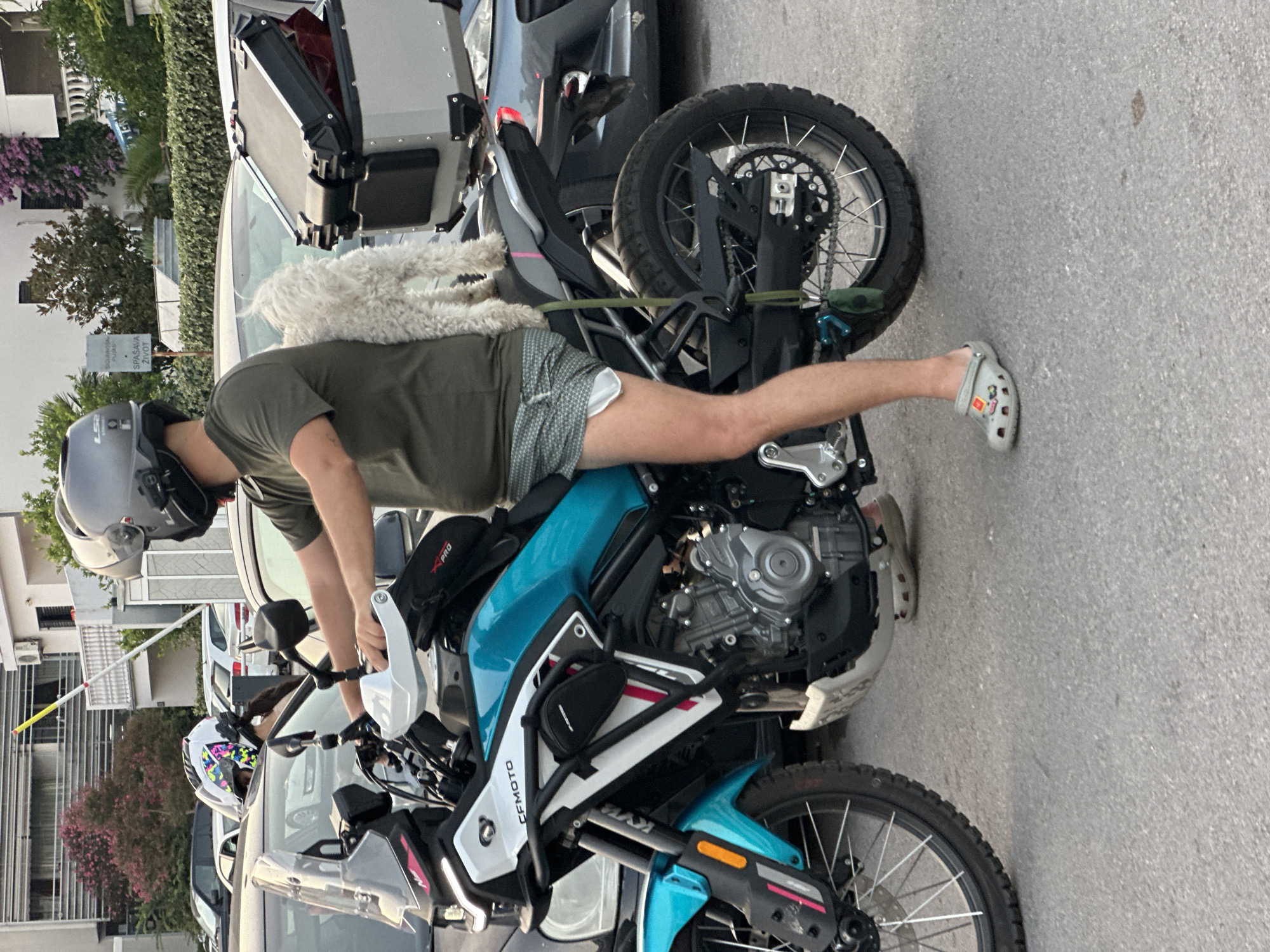 A person wearing a helmet, olive green t-shirt, and patterned shorts is sitting on a turquoise and white motorcycle. A white, fluffy dog is standing on the seat behind the person, with its front paws resting on the person's shoulders. The motorcycle has saddlebags on the rear and a windshield. The person's left foot is visible, wearing a grey Croc sandal with charms. The scene appears to be on a paved street in Budva, Montenegro, with other vehicles parked in the background and buildings and foliage visible.