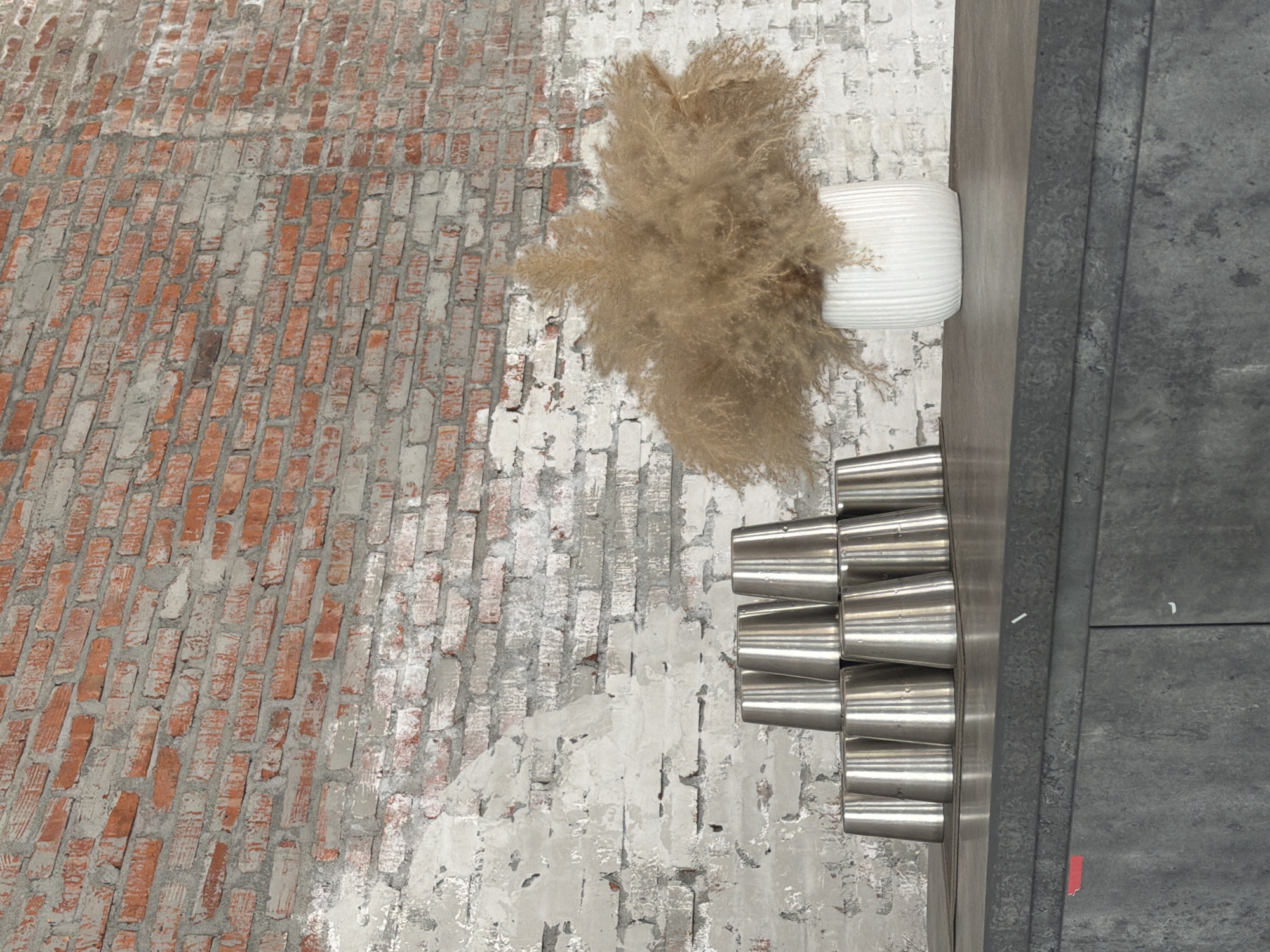 A high-angle shot shows a partially whitewashed brick wall on the left transitioning to a more uniformly whitewashed wall with exposed brick remnants. A white cylindrical vase holding dried foliage sits atop the wall. To the right of the vase, a shelf or counter supports a stack of silver, cone-shaped metal objects. Further right, a dark gray surface with a layered appearance extends to the edge of the frame. The scene lacks people, actions, and interactions. There is no definitive information to confirm that this is a scene in Đà Nẵng, Vietnam.
