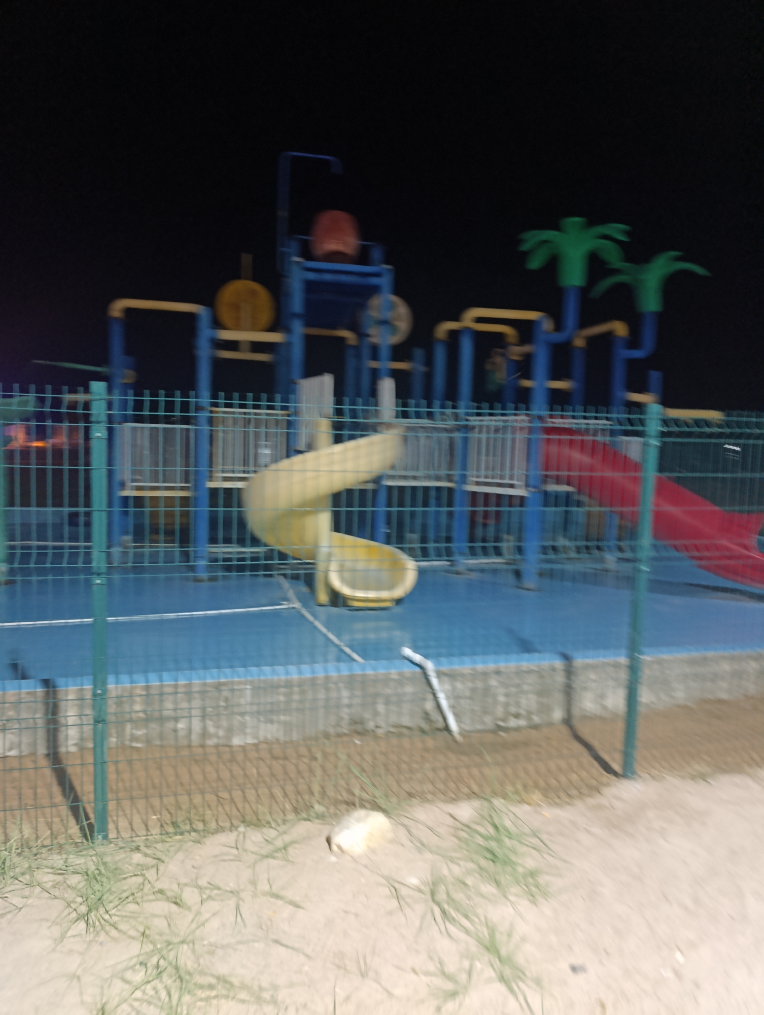 This is a nighttime photograph of a playground structure in Bosteri, Kyrgyzstan. The structure is multi-colored, featuring blue posts, yellow railings, and red and yellow slides. Two green palm tree-like decorations are visible at the top. The playground is enclosed by a green metal fence. The ground inside the fence is blue, and the area outside appears to be sand with some sparse grass. The scene is dimly lit, suggesting it is either late at night or in an area with limited artificial lighting. There are no people or animals visible in the image. The image is slightly blurry, possibly due to low light conditions and camera movement.