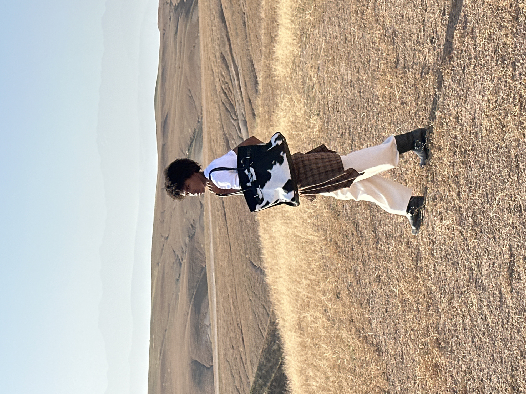 The image shows a person standing in a field with mountains in the background. The person is wearing a white t-shirt, light-colored pants, and black boots, and has a patterned tote bag. The location appears to be a dry, grassy area. While the image shares landscape characteristics with Sidi Badhaj, Morocco, there is no way to confirm that the scene is in that specific city from visual evidence alone. The person is facing to the left. The ground is covered in dry grass. The sky is clear and blue.
