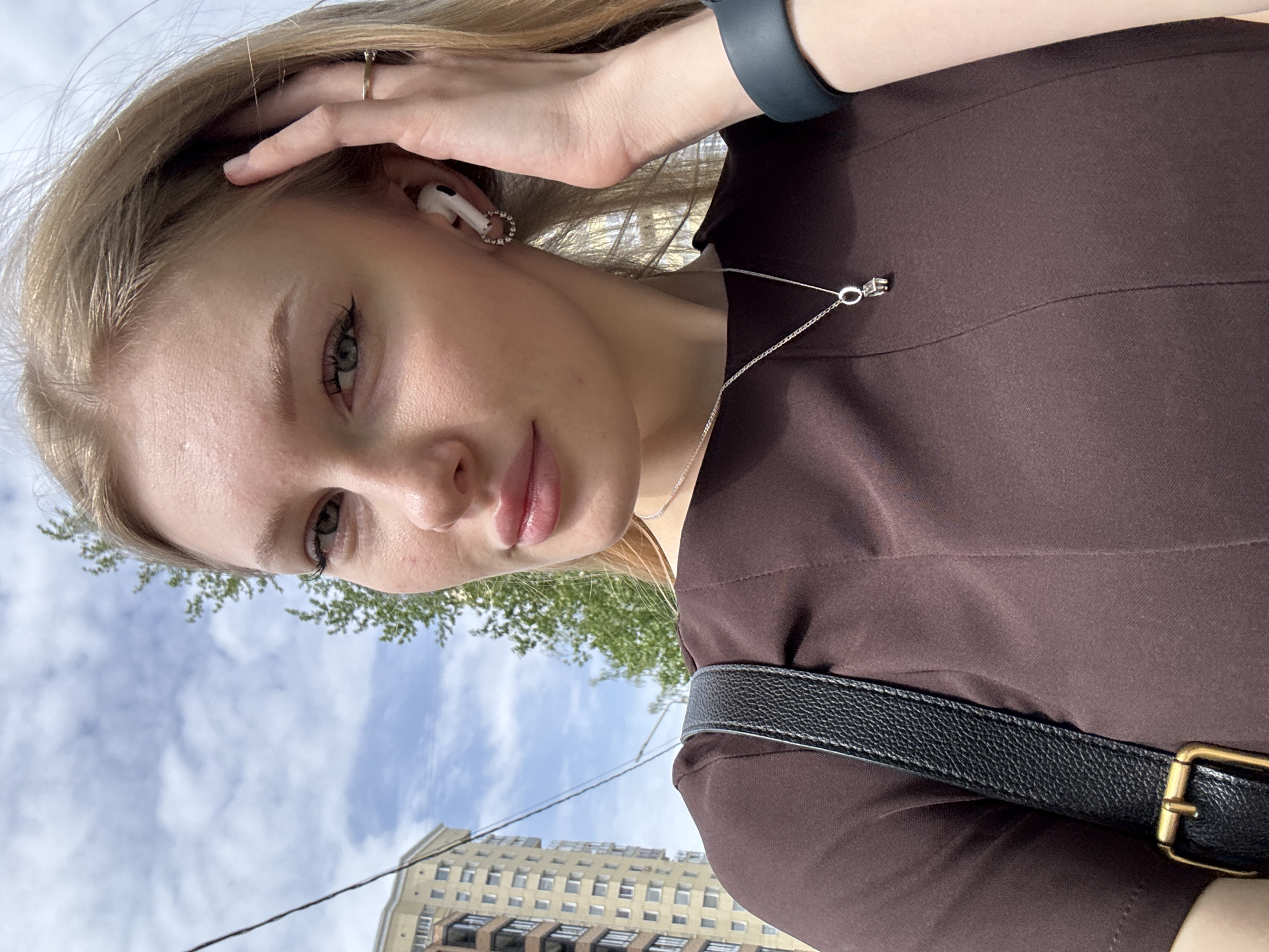 A selfie of a young Caucasian woman with fair skin and blonde hair, taken outdoors during the day. She is wearing a brown dress, a silver necklace, and a dark-colored smartwatch. She has a ring on her finger and an earpiece in her ear. Her left hand is partially raised, touching her hair. The background includes a blue sky with clouds, green tree foliage, a multi-story building, and a visible powerline. The buildings indicate that the location is likely within the city of Perm, Russia. A black strap with a buckle is visible across the dress, potentially a purse strap.