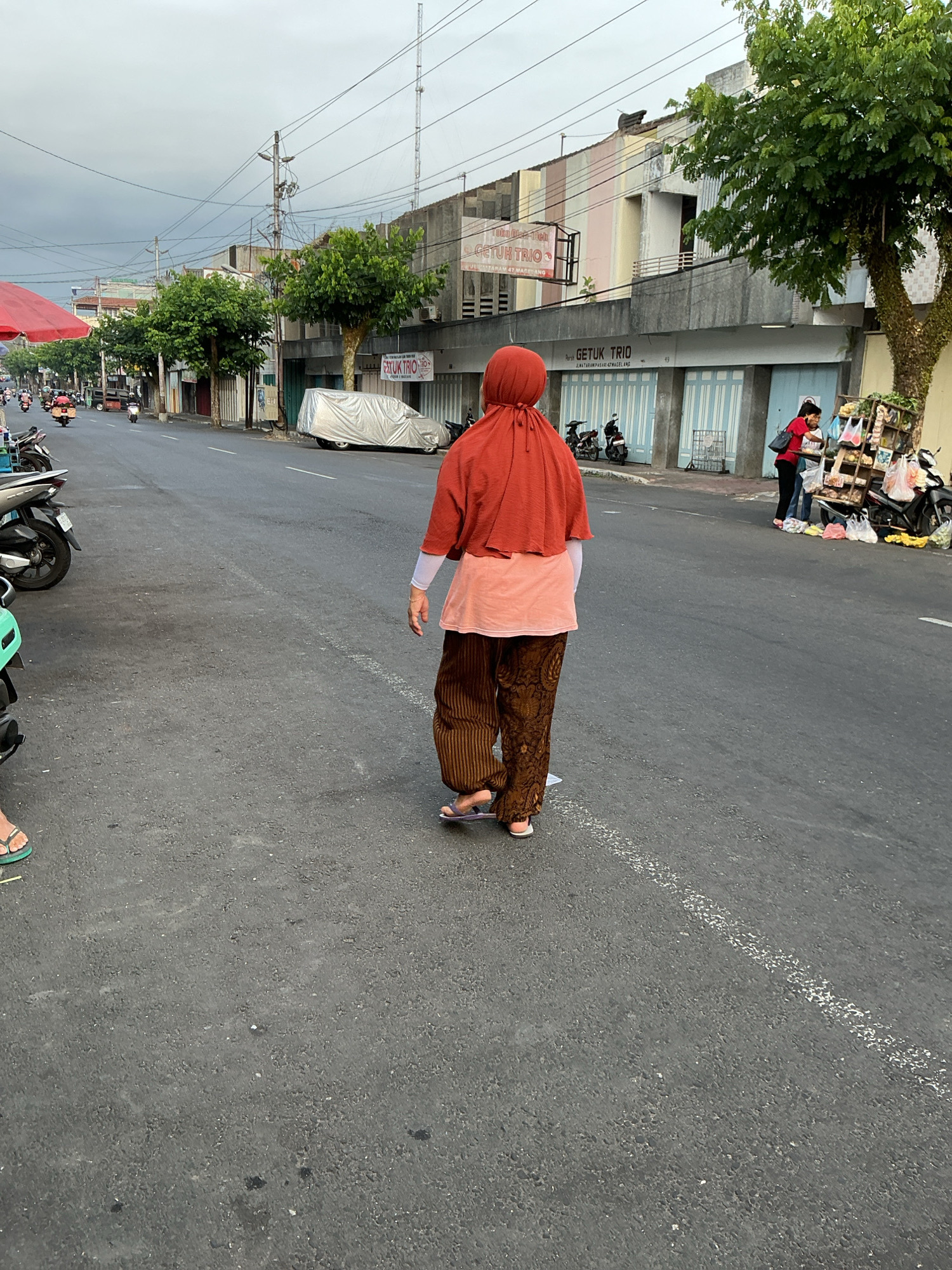 An outdoor, eye-level shot captures a street scene in Magelang, Indonesia, characterized by a cloudy sky and overcast lighting. In the foreground, a woman wearing a rust-colored hijab and a light pink blouse walks away from the camera, her patterned brown pants and flip-flops visible. The street is asphalt, marked by a white line. To the left, parked motorcycles line the side of the road, and a red awning extends from a stall. Further down the street, a silver car is covered with a tarp, and a vendor stands behind a cart displaying various goods, attended by another person in a red shirt. Buildings with pastel-colored stripes line the street, and trees with green foliage add to the urban landscape. Power lines crisscross the sky, and a tall antenna is visible in the distance.