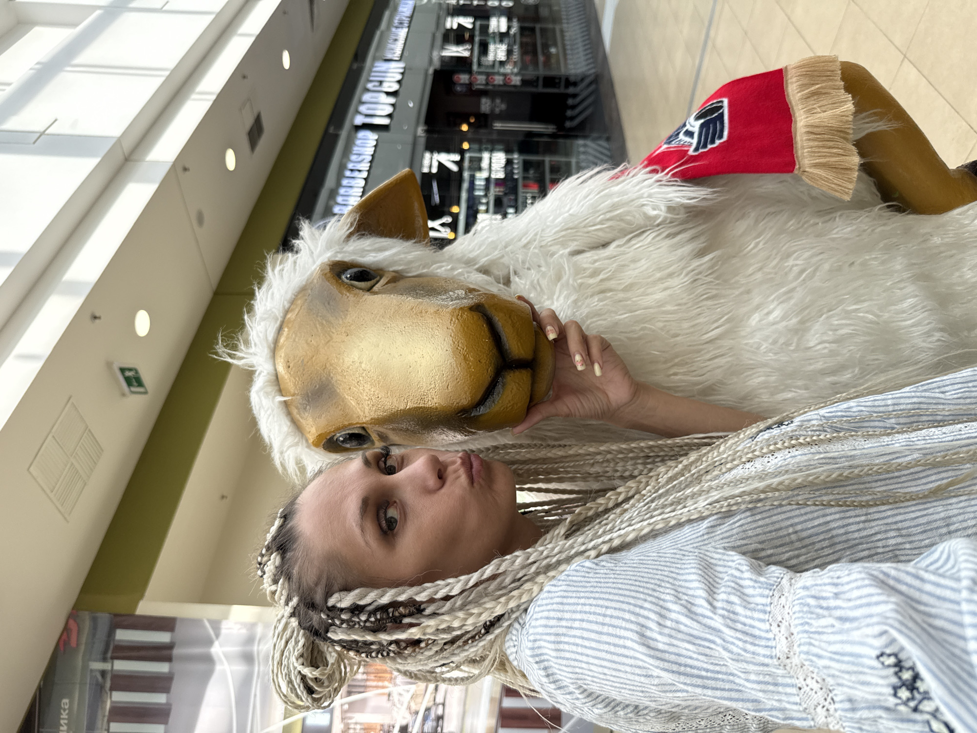 A woman with blonde braids is taking a selfie with a mascot costume of a sheep. The sheep costume has a golden face, white fluffy fur, and a red arm band with a blue logo. The woman is wearing a blue and white striped shirt and has her lips pursed. In the background, there are shops, including a barbershop called "TOP GUN". The scene appears to be indoors, likely in a shopping mall.