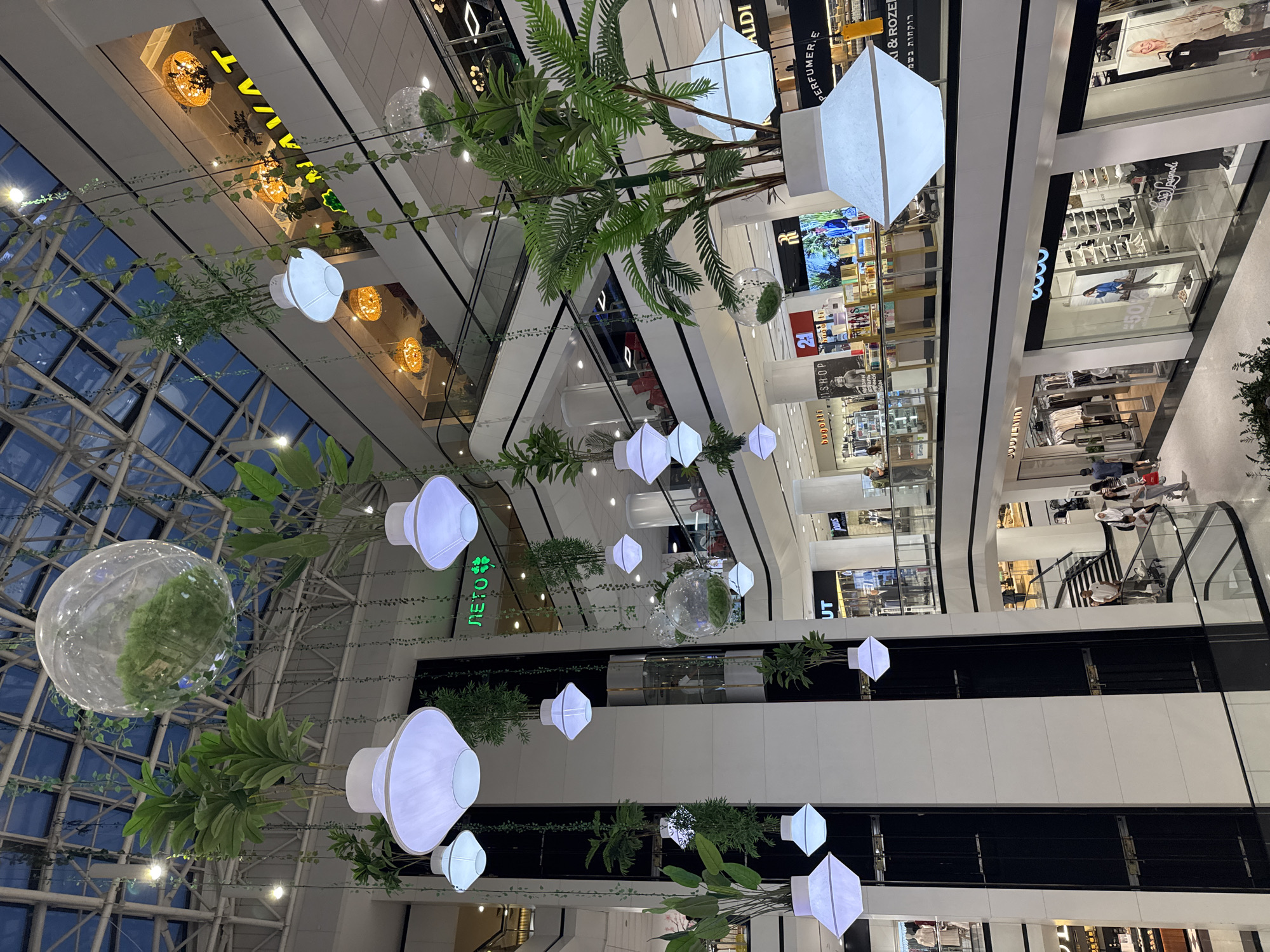 The image depicts the interior of a multi-level shopping mall with a glass ceiling. Hanging from the ceiling are numerous white, geometric pendant lights and transparent spherical glass containers holding greenery. Plants are also growing from large white planters, with vines cascading down. In the background, escalators and various store fronts are visible across different levels, including shops with signage such as "BUGOTTI," "SHOP," "21," "RI," "ALDI," and "PERFUMERIE." Several people are present, some walking on the mall's upper walkways and escalators, while others are browsing in the stores. The overall atmosphere is bright and modern, suggesting a commercial space designed with a natural, airy aesthetic.