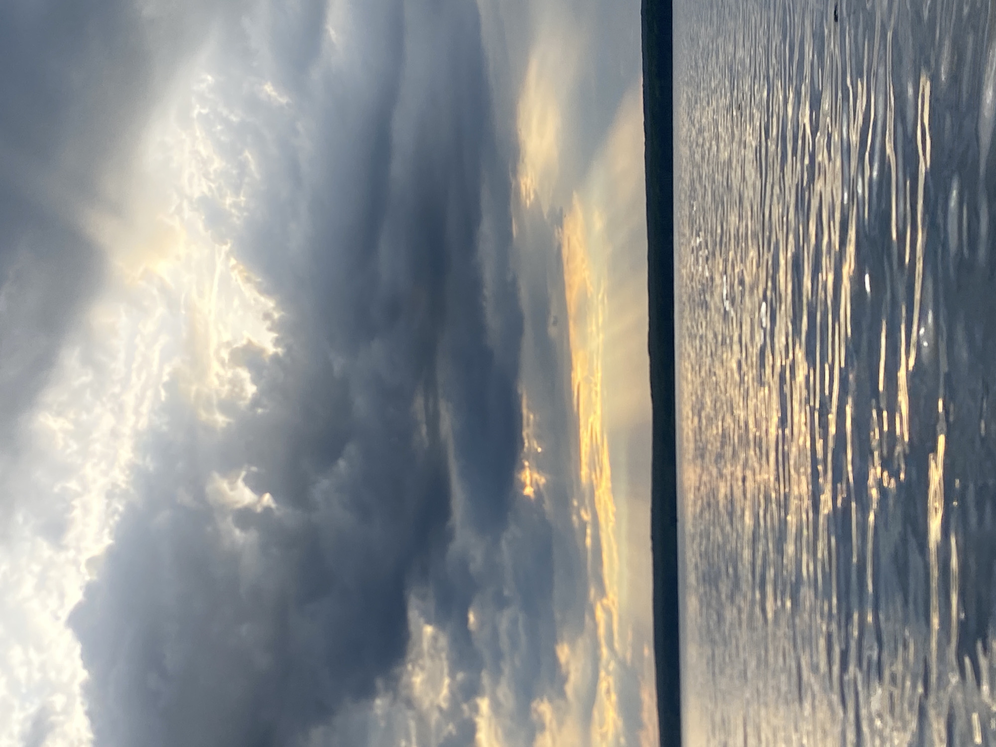 The image presents a tranquil natural landscape, capturing a dramatic sky above a body of water, likely a river or lake, near Chaikovsky, Russia. The scene appears to be set during late afternoon or early evening, possibly at sunset, indicated by the warm, golden light.
The upper portion of the image is dominated by an expansive sky filled with dynamic cloud formations. Dark, dense clouds are interspersed with areas where bright, golden sunlight penetrates through, forming distinct crepuscular rays. These sunbeams create a striking visual contrast, highlighting the textures and depths of the clouds and casting a luminous glow across parts of the sky.
Below the horizon, a calm body of water stretches across the frame. Its surface is covered with numerous small ripples and waves, which brilliantly reflect the golden light from the sky. These reflections appear as shimmering, elongated streaks on the water, adding a sense of movement and enhancing the serene atmosphere. The horizon itself is a subtle, dark line separating the luminous sky from the reflective water.
There are no people, identifiable man-made objects, or visible text in the photograph. The focus is entirely on the natural elements and the interplay of light and clouds over the water, conveying a sense of peaceful grandeur and the beauty of dusk.