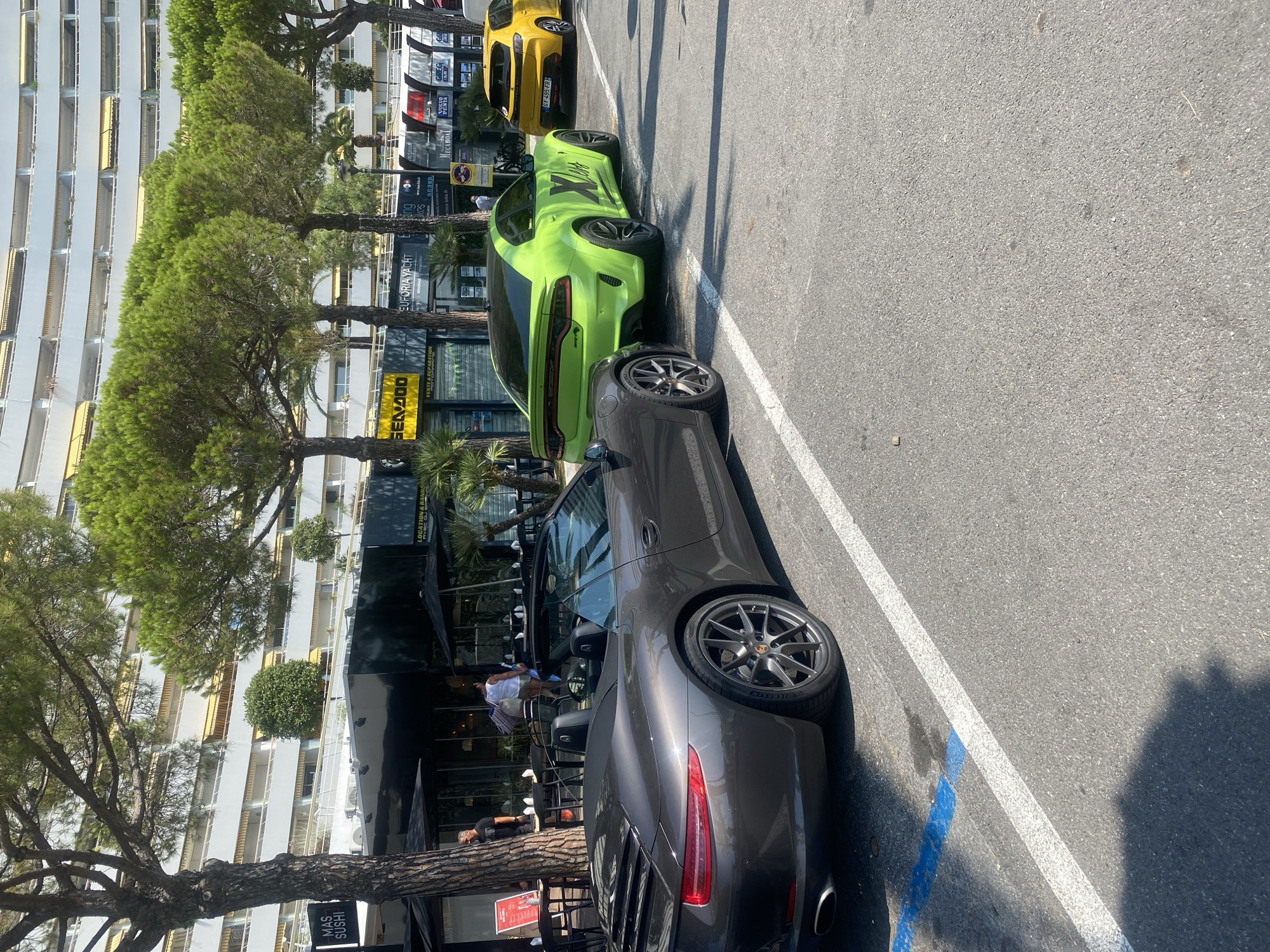 In Villeneuve-Loubet, France, three cars are parked in a row on a sunny day. The closest vehicle is a dark gray convertible sports car. To its right is a bright green sedan with racing stripes. The furthest car is a yellow hatchback. In the background, a modern building with multiple balconies and yellow awnings stands behind a line of pine trees. A sidewalk and shops are visible in front of the building.
