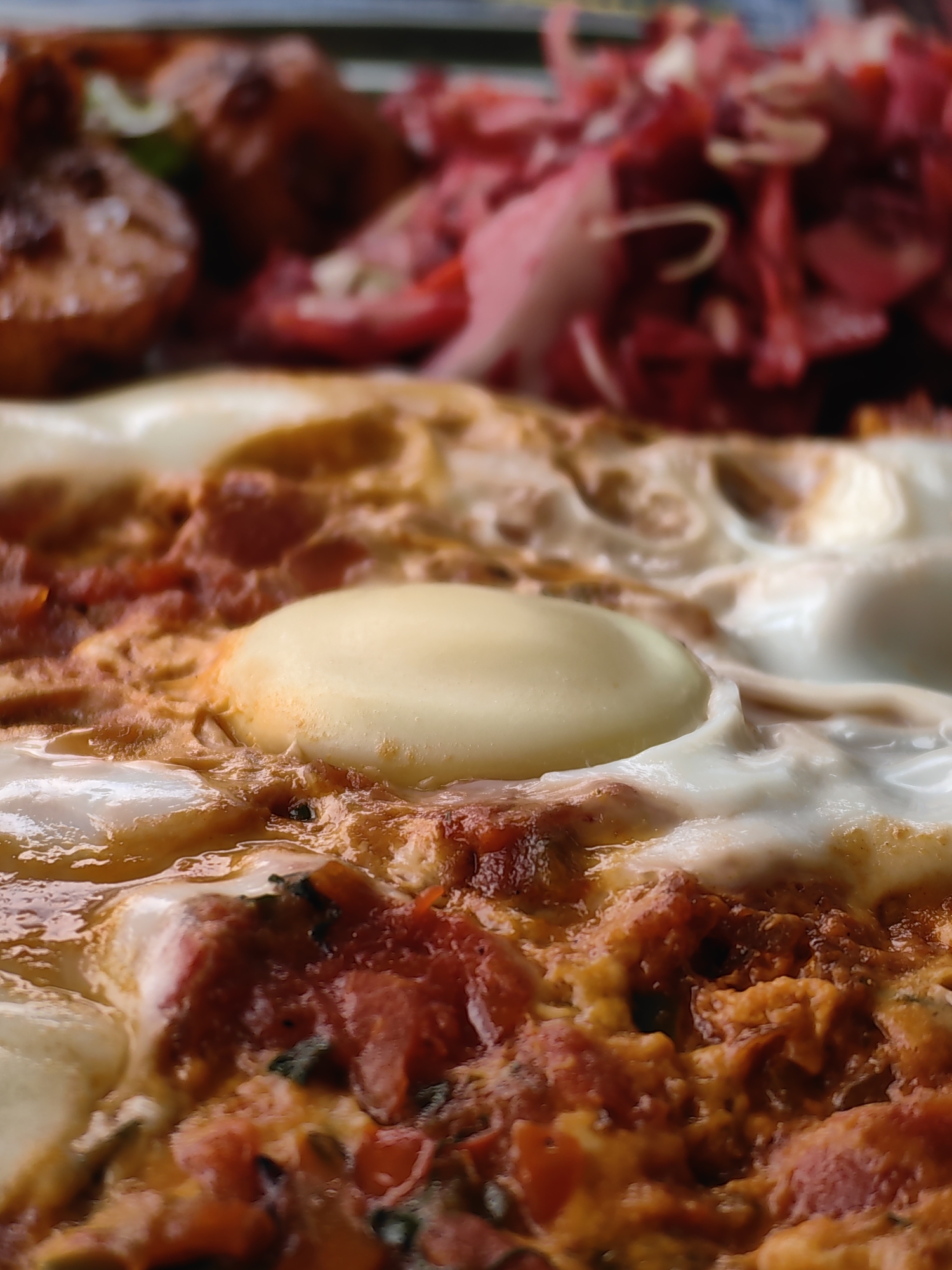 The image displays a close-up view of a dish, likely a breakfast item, featuring eggs cooked in a tomato-based sauce. The eggs, with their distinct white and yellow portions, are nestled within the savory mixture. In the blurred background, elements of a meal are discernible, including what appears to be some kind of meat or protein dish on the left and a vibrant red salad, possibly made of cabbage and onions, on the right. The setting suggests a meal in Vashisht, India, an area known for its hot springs and spiritual retreats, implying a relaxed and possibly traditional culinary experience.