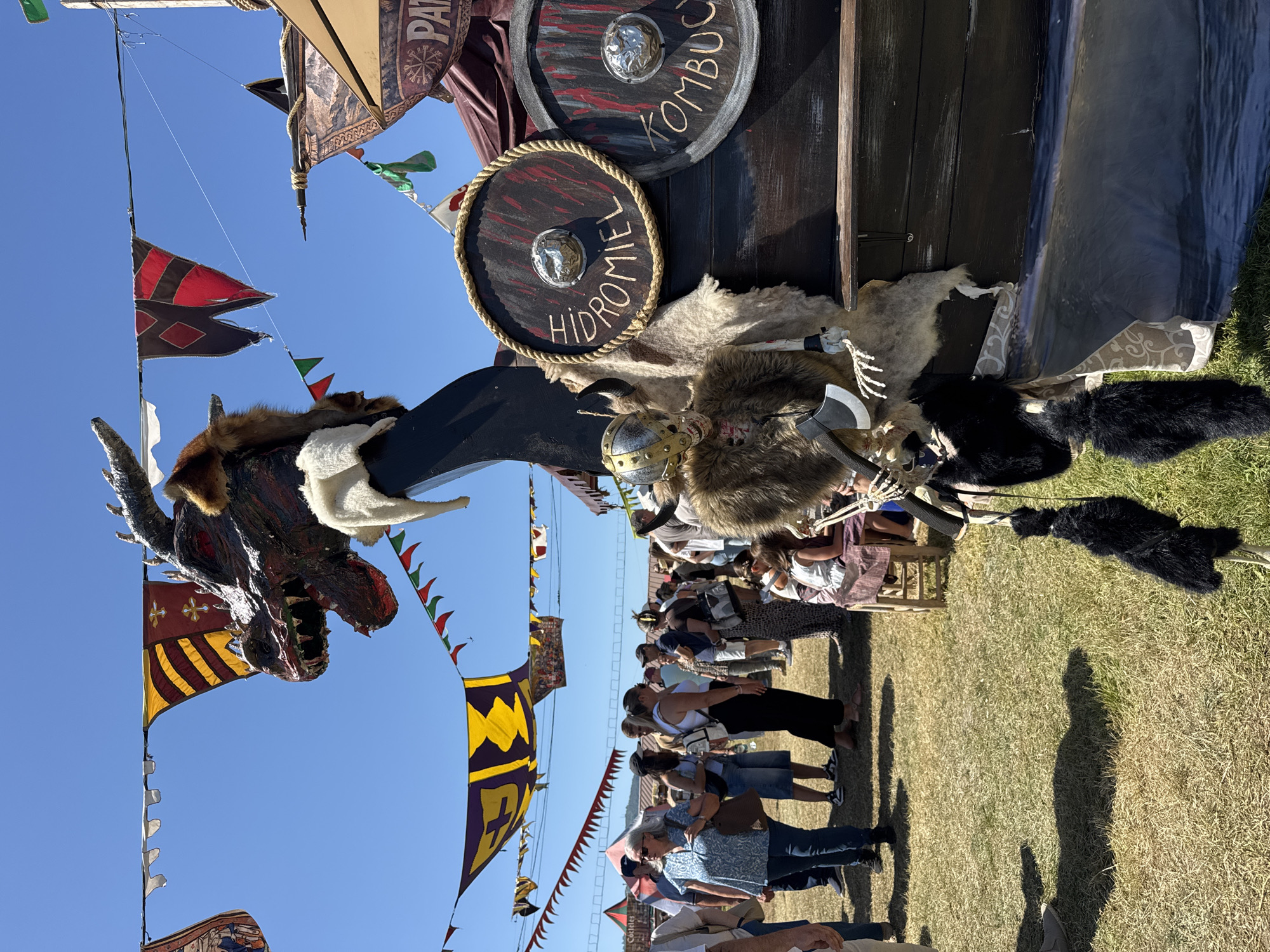 The image captures a vibrant outdoor event, likely a medieval fair, held under a clear blue sky. In the foreground, a section of a Viking ship's prow is visible, adorned with a fearsome dragon head figure with sharp teeth and red eyes. A skeletal figure dressed in a fur cloak and a helmet, carrying an axe, is positioned beside the ship. Two round shields are attached to the ship's side; one is labeled "HIDROMIEL" and the other "KOMB", presumably indicating mead and kombucha. Hanging from the ship are decorative black furry elements. In the background, a crowd of people are mingling, dressed in casual modern clothing, with some wearing hats. Colorful banners are strung across the sky, adding to the festive atmosphere. The ground is covered with dry grass, suggesting a warm climate. A tent with the word "COCTELES" is visible in the far left background. This scene is consistent with events like the "Mercado Medieval" held annually in the city of Llanera, Spain, known for its historical reenactments and cultural festivals.