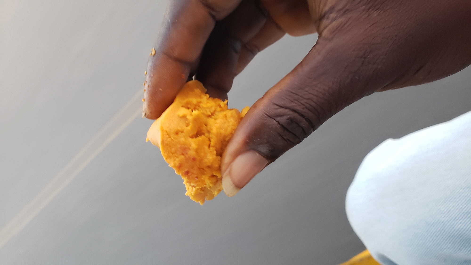 A person is holding a piece of bright orange food, possibly cornbread or a sweet potato-based pastry, between their thumb and index finger. The food is roughly spherical with an irregular surface texture. The person's hand is dark-skinned, with visible wrinkles and a short, light-colored fingernail on the index finger. They are wearing light blue jeans. The background is a blurry, light gray, suggesting an outdoor setting with a neutral or overcast sky. The image is taken from a close-up perspective, focusing on the hand and the food. The context provided about Jalingo, Nigeria, is not directly observable from the visual elements of the image itself.