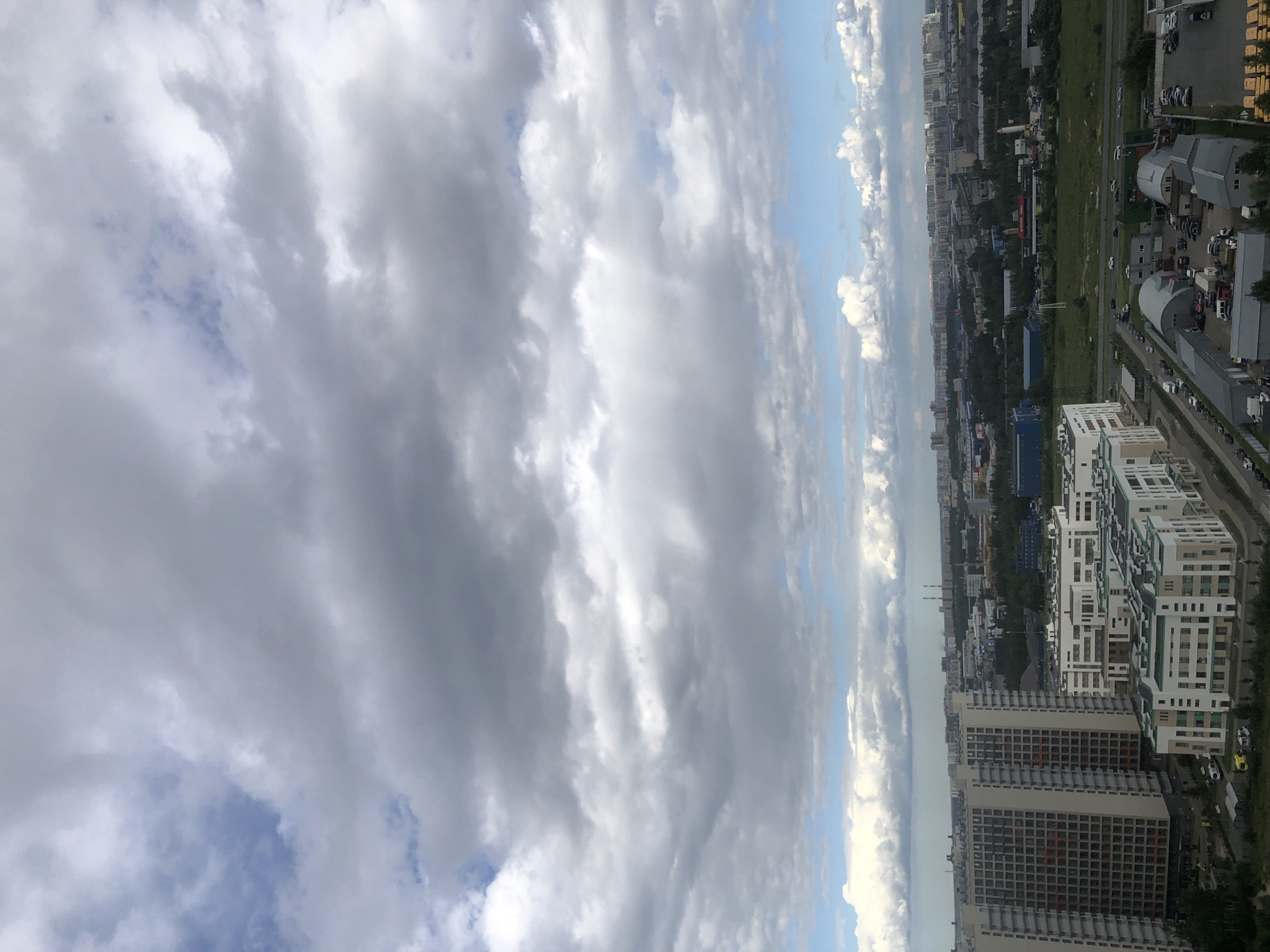 This is a photograph taken from a high vantage point, looking down on an urban landscape under a cloudy sky. The image is oriented vertically, with the sky occupying the left side of the frame and the cityscape stretching across the right.
The sky is filled with a dramatic display of clouds. Some are dark and heavy, suggesting impending rain, while others are bright and white, allowing sunlight to peek through. Patches of blue sky are visible between the cloud formations.
The cityscape below consists of a dense collection of buildings, primarily residential high-rises. These buildings exhibit a variety of architectural styles, with some featuring modern, geometric designs and others appearing older and more uniform. Green spaces, likely parks or undeveloped land, are interspersed between the structures. A road or highway runs through the scene, with a line of cars visible. Some industrial buildings and infrastructure, like smokestacks, can also be spotted in the distance.
There is no discernible activity or event taking place in the image, suggesting it is a still observation of the urban environment. The time of day appears to be daytime, given the lighting conditions. The weather seems changeable, with both cloudy and potentially sunny elements. No specific location cues are obvious, and no emotions are conveyed. No visible text is present in the image.