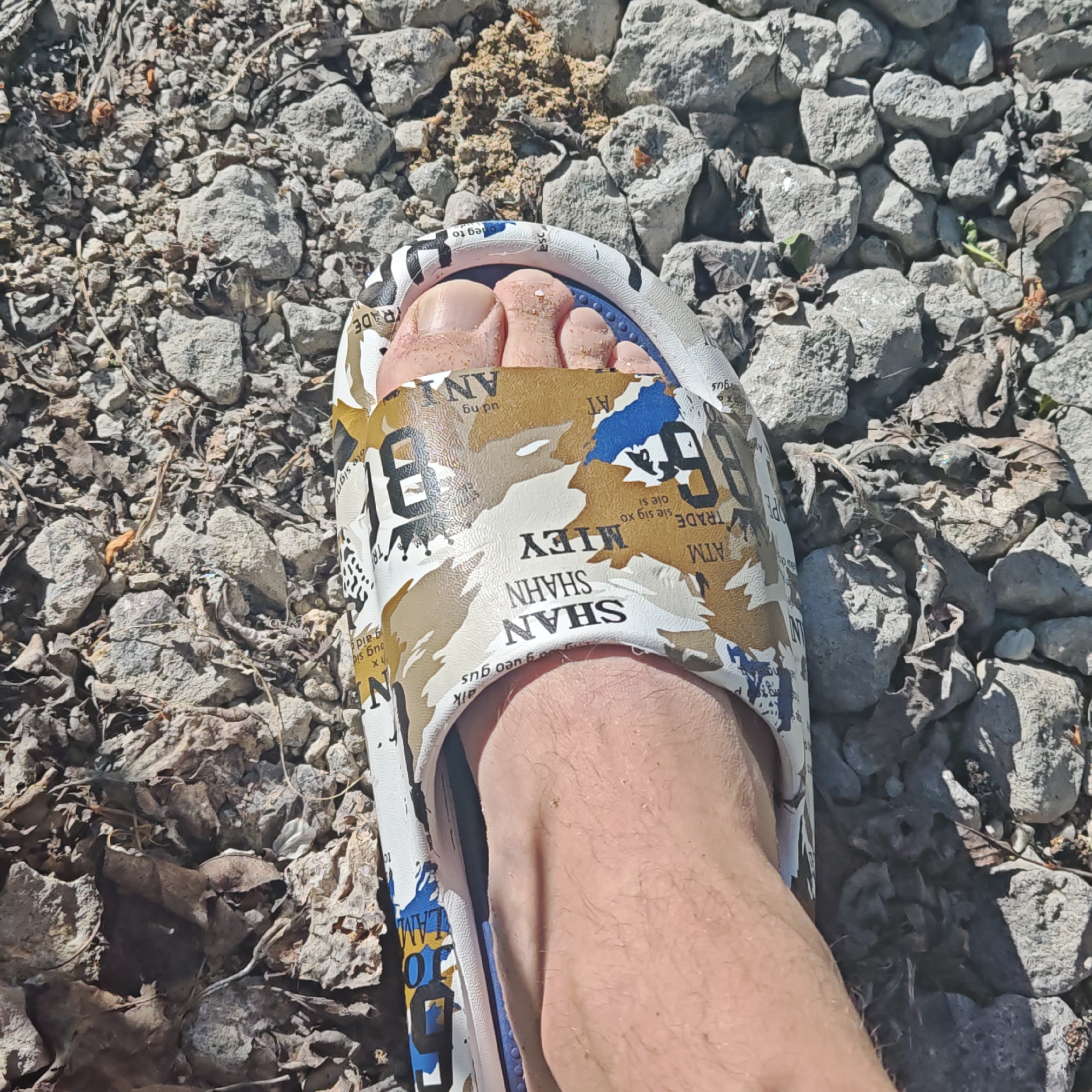 The image shows a close-up of a person's foot wearing a white sandal with a colorful, urban-themed design. The sandal is resting on a surface covered with small, gray rocks and some dried leaves. The environment appears to be outdoors, possibly a pathway or ground area covered with gravel. The sandal design includes words like "SHAN," "SHAHN," "MIEY," "ATM," "TRADE," "ANI," and "LAM" among other elements. The location in the city of Unknown, Unknown cannot be determined based on this image.