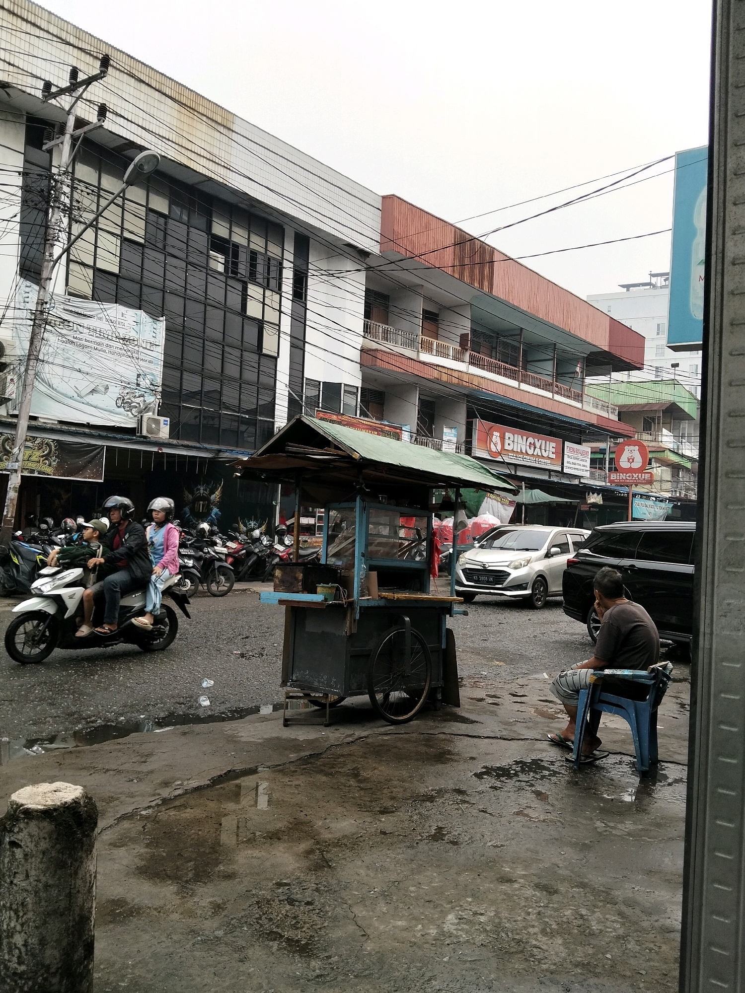 The image depicts a street scene in Pontianak, Indonesia, likely a commercial area. A multi-story building with a dark glass facade dominates the background. In the foreground, a food cart with a green canopy is positioned on a wet asphalt road. Several motorcycles are parked to the left of the cart, and two people are riding a white scooter in motion. To the right of the food cart, a man is seated on a blue plastic chair, facing away from the camera. A black SUV is parked behind him. The sky is overcast, and the overall atmosphere suggests a humid or recently rained-on environment. Several signs are visible on the buildings, including one that reads "BINGXUE".