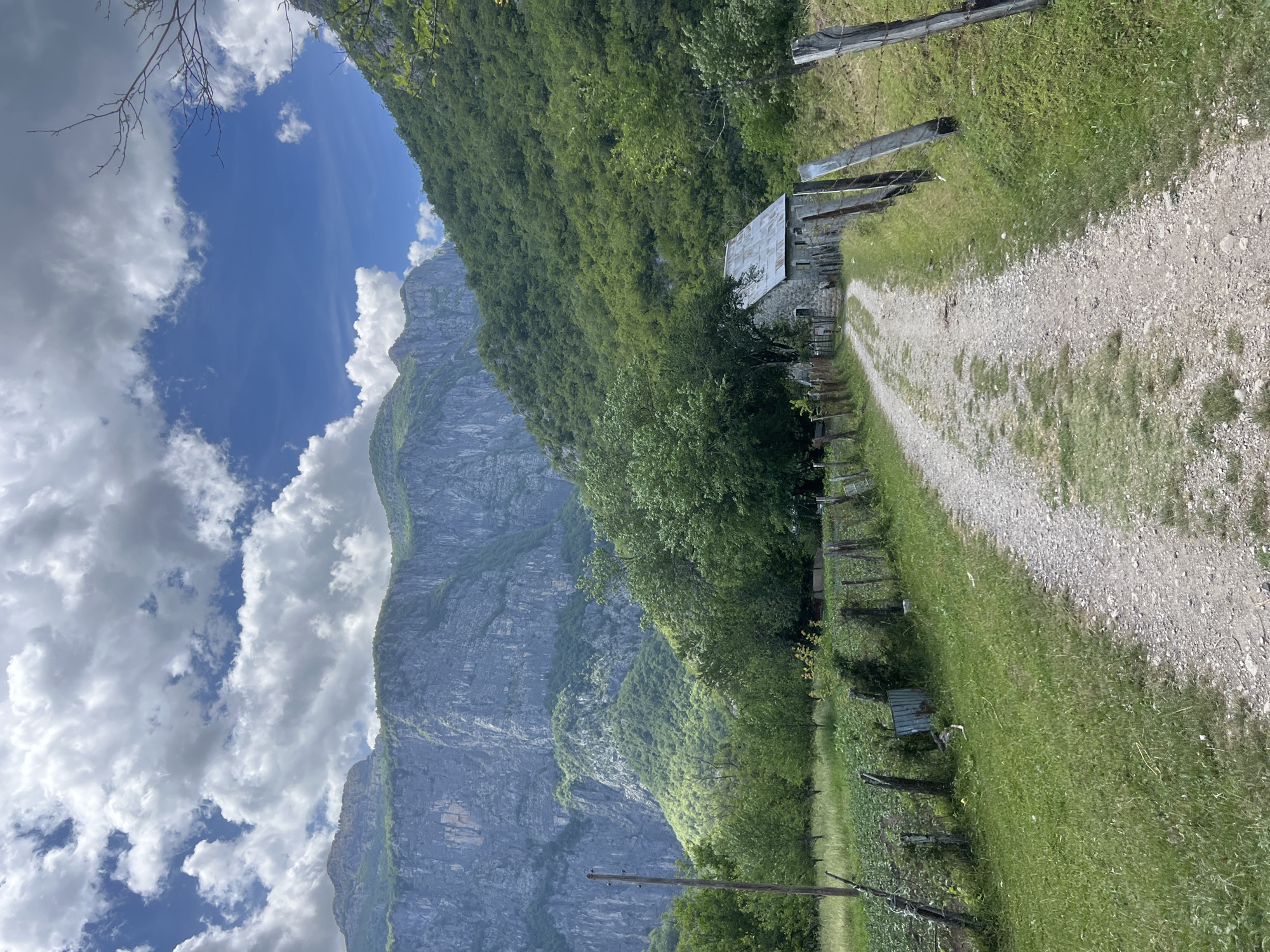The image captures a rural landscape in Mrtvo Duboko, Montenegro. A gravel path leads into the scene, flanked by grassy verges and a rustic wooden fence on one side. A small, stone building with a metal roof is situated along the path, partially obscured by trees. A steep, rocky mountain dominates the left side of the frame, contrasting with a sky filled with blue patches and puffy white clouds. A few power poles are visible along the lower portion of the image.