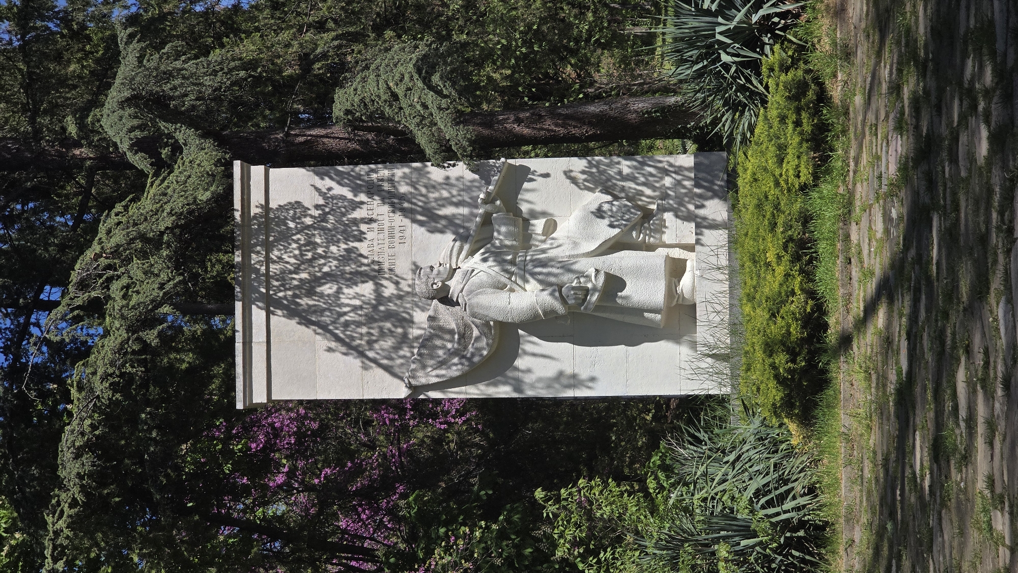 The image shows a stone monument of a soldier in the city of Stara Zagora, Bulgaria. The monument is set against a backdrop of trees and shrubbery. The soldier figure is depicted in relief, and text in Cyrillic script appears on the monument's surface. The surrounding environment consists of a mix of greenery, including trees, bushes, and possibly some flowering plants, with dappled shadows cast on the monument and the adjacent ground. The ground appears to be paved with stone blocks or slabs with some grass.