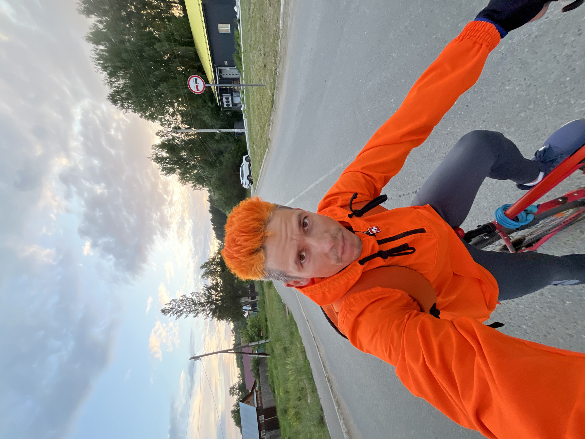 A man with bright orange hair is riding a bicycle on a paved road in Chaikovsky, Russia. He is wearing an orange hooded jacket and gray leggings. The sky is partly cloudy, with hints of sunset colors. In the background, there are trees, houses, and a white SUV. A traffic sign indicating no overtaking is visible on the right side of the road. The man is looking directly at the camera, appearing to be taking a selfie.