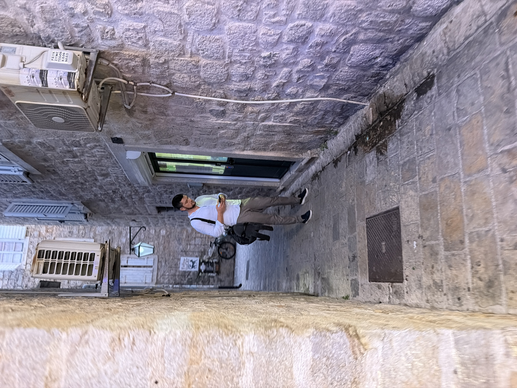A man stands in a narrow cobblestone alley in Budva, Montenegro, looking at his phone. He is wearing a white shirt and grey pants, and has a black backpack slung over his shoulder. The alley walls are made of stone, and there is an air conditioning unit mounted on one of the walls. A bicycle is parked further down the alley. The scene is dimly lit, suggesting it is either evening or a shaded area.