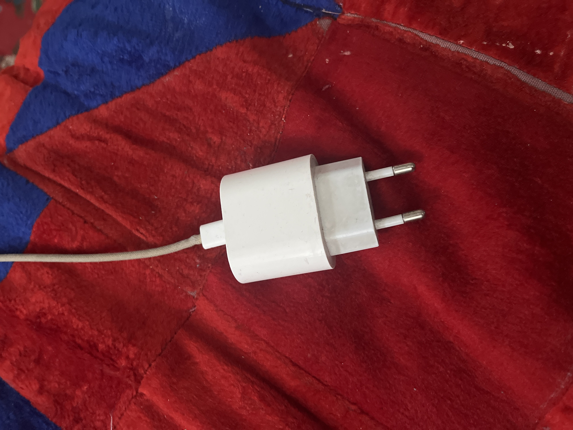 A white USB charger adapter with two prongs is positioned on a red and blue fuzzy fabric. A thin, beige cable extends from the top of the adapter and disappears out of the frame. The fabric appears to be a blanket or throw, with a visible seam running diagonally across the image. The lighting suggests an indoor setting. The image was taken in Boraldai, Kazakhstan.