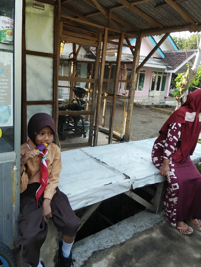 This image, taken in Pesawahan, Indonesia, depicts an outdoor scene with two individuals and multiple structures under clear daylight conditions. In the left foreground, a young person, possibly female, wears a brown scout uniform with a red and white neckerchief, a dark brown hijab, brown trousers, white socks, and black shoes. She is seated on a low concrete ledge, holding a multi-colored popsicle or candy to her mouth and looking slightly towards the viewer. To her right, a makeshift sales counter or bench covered with a white tarp is visible, supported by wooden beams, with a dark, open area beneath it. Seated on this counter, facing away from the camera and towards the right, is an adult female wearing a dark red hijab, a long-sleeved dark red dress with white patterned accents, and orange flip-flops. Her right hand rests on the edge of the counter.
Behind the individuals and the counter is a wooden framework structure, possibly part of a stall or enclosure, with wire mesh panels and a corrugated metal roof. Inside this structure, indistinct machinery, possibly a grinder, is visible on a stand. In the background, beyond the wooden structure, stands a light pink house with white trim, blue accents on its gabled roof, and a brown tiled roof. Green foliage is visible to the right of the house. The immediate ground in the foreground is concrete, showing some discoloration. To the far left, a silver-framed display unit with text in Indonesian, including "TIGA SA" and items like "latte," is partially visible.