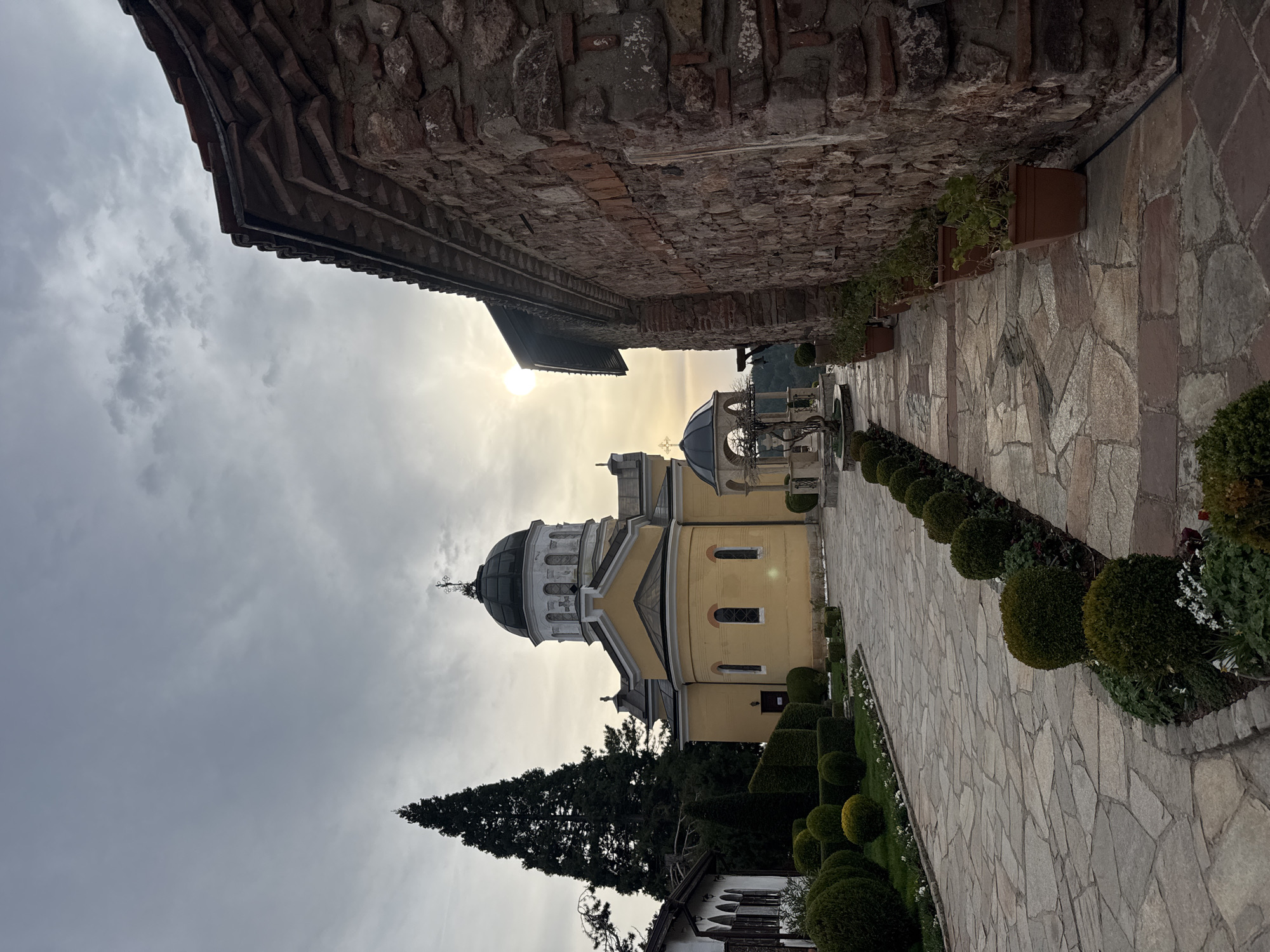 The image depicts a church with a yellow facade and a dark dome, located in Sofia, Bulgaria. The church is viewed from a stone pathway lined with meticulously trimmed, round shrubs and some taller, square bushes. To the right, a high stone wall runs parallel to the path, featuring potted plants affixed to its surface. A small structure with a domed roof stands behind the church. The sky is overcast with the sun trying to break through the clouds. The image appears to have been taken at ground level.