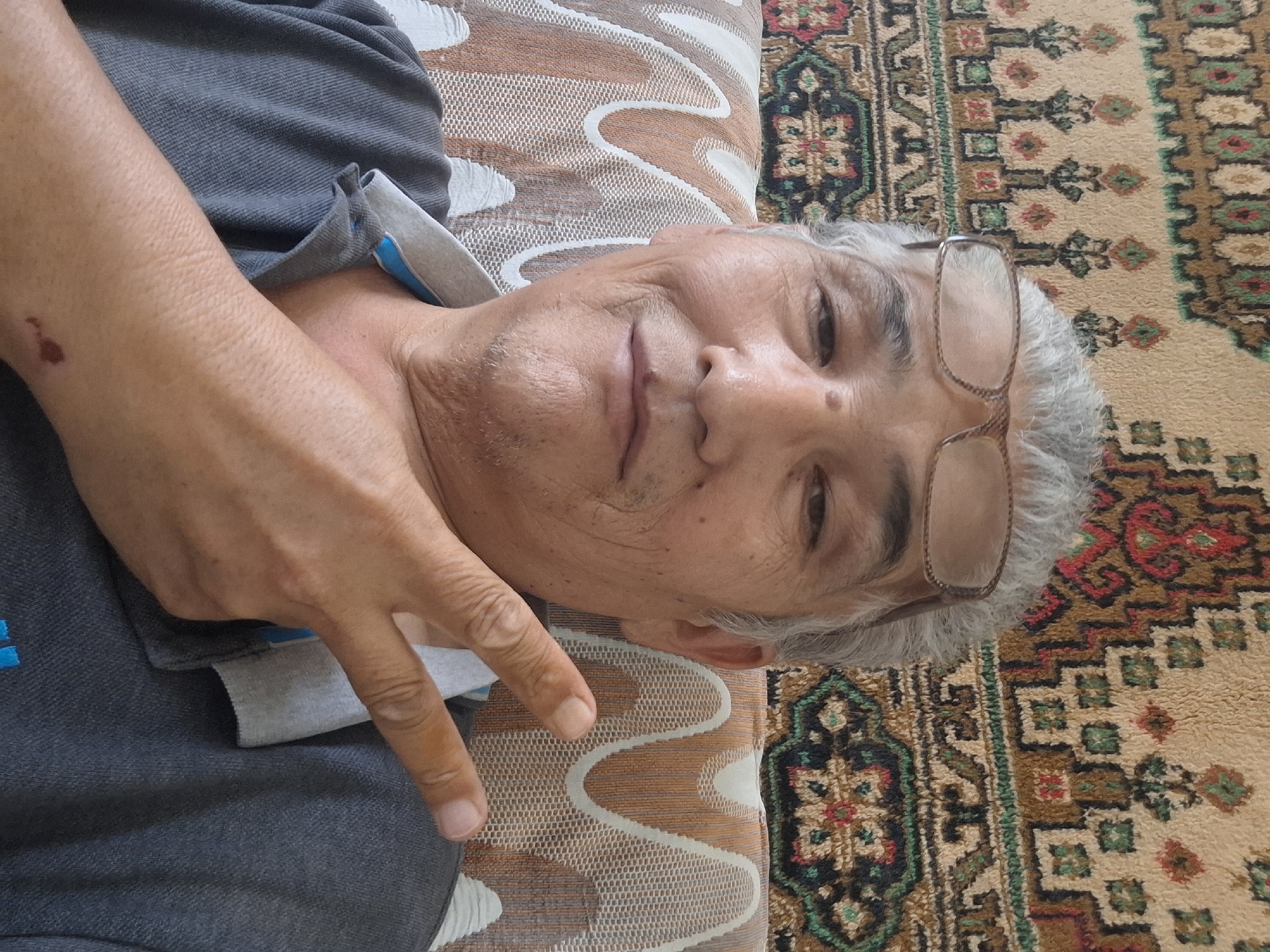 A middle-aged man with gray hair and glasses is taking a selfie in what appears to be a living room in Almaty, Kazakhstan. He is wearing a gray polo shirt with a blue collar. He is making a peace sign with his right hand. The background shows a patterned carpet and a couch. The man's face is wrinkled, and he has a mole on his cheek. There is a small wound on his left arm. The lighting suggests the photo was taken during the day.