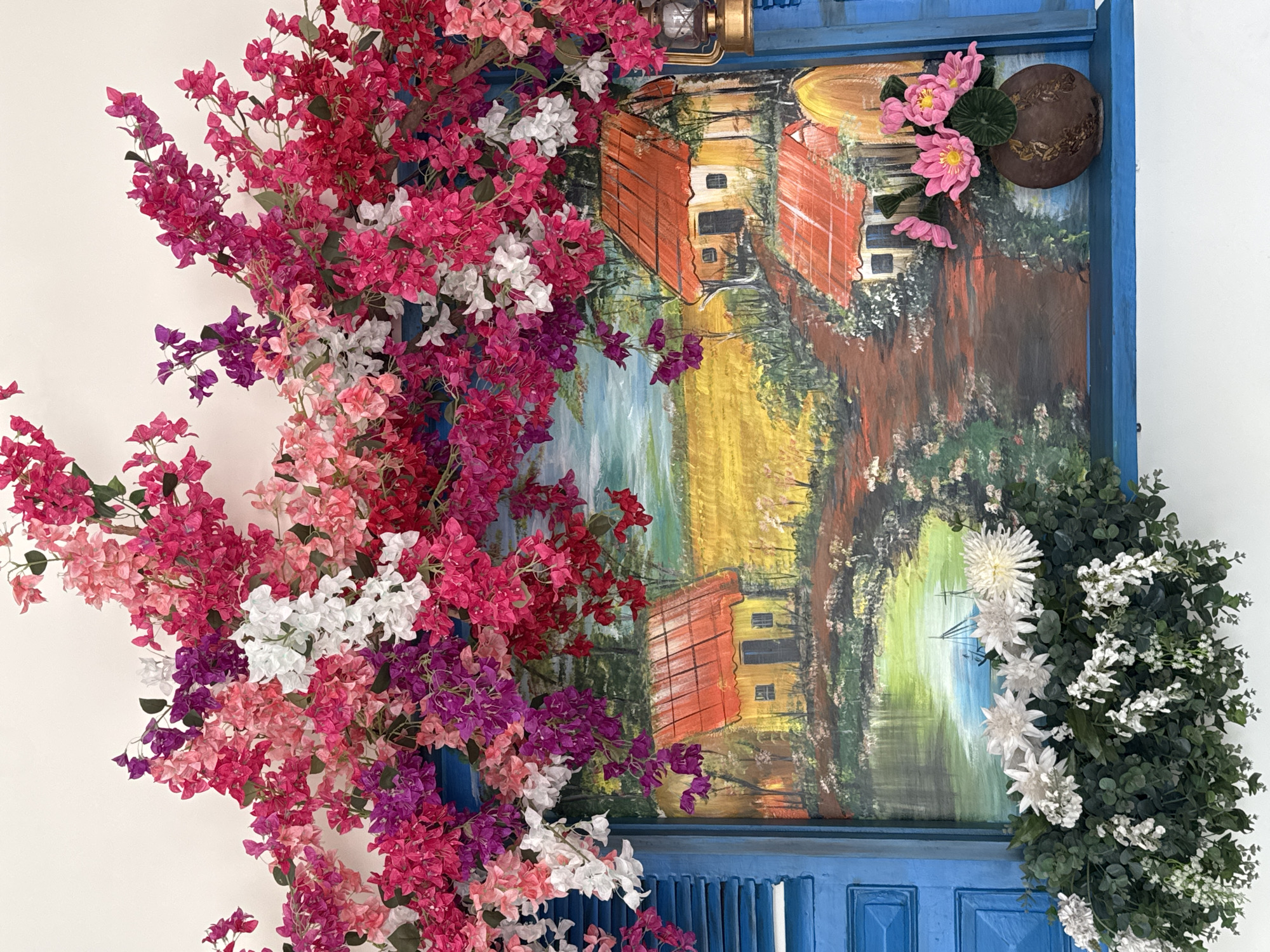 The image depicts a blue-framed painting of a rural Vietnamese landscape, adorned with vibrant, colorful artificial flowers. The painting features a scene with houses that have orange tile roofs, possibly near water or a river. The frame itself appears to be made to resemble a shuttered window. The floral arrangement includes a mix of pink, purple, red, and white bougainvillea. A lantern sits on top of the frame. A brown pot with attached pink flowers sits on the upper right of the artwork. The overall aesthetic suggests a decorative element of a business in Đà Nẵng, Vietnam that aims to capture a traditional or artistic theme. There are no people visible in the image.